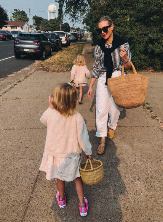 Karin Emily walks with her kids on a sidewalk and wears a black and white striped long sleeve tee white baggy jeans from agolde, nude fisherman sandals, a basket bag, and a navy cashmere sweater over her shoulders