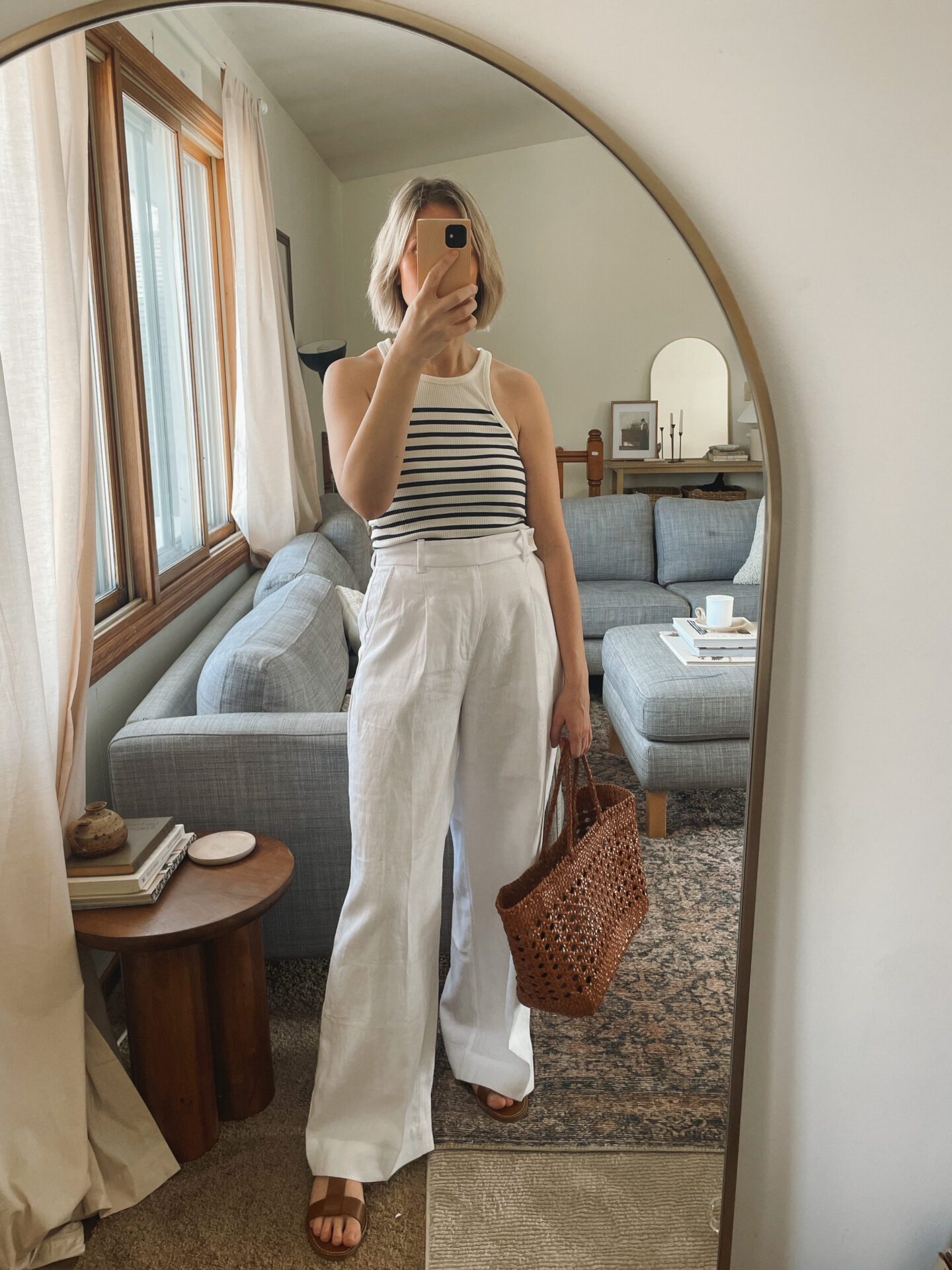 Karin Emily takes a mirror selfie in her living room and wears a striped tank top and white linen pleated wide leg pants with a dragon diffusion bag