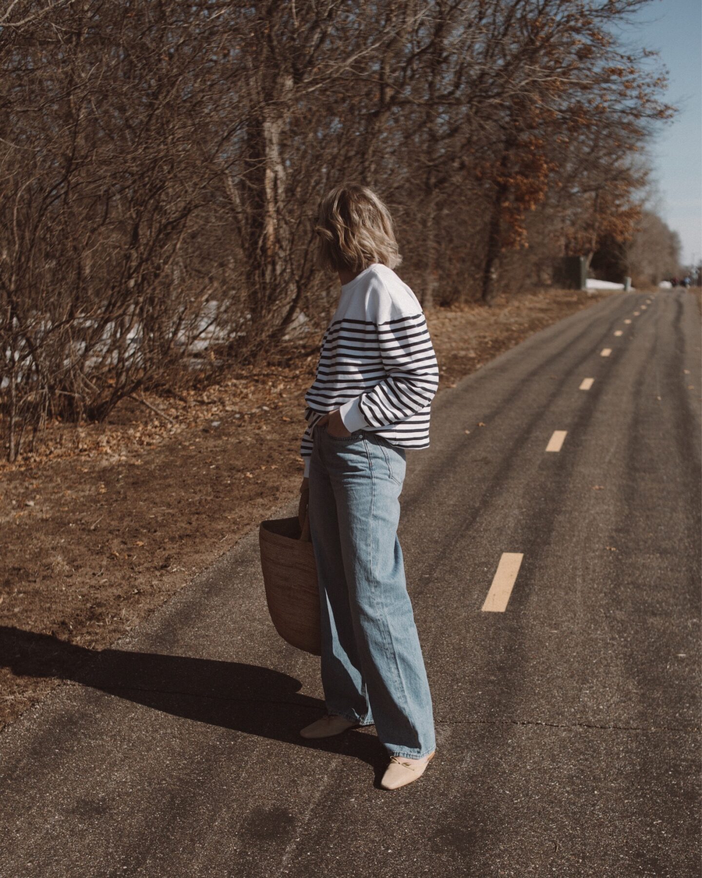 Karin Emily stands on a path and wears a stripe sweatshirt, baggy jeans and nude colored mules