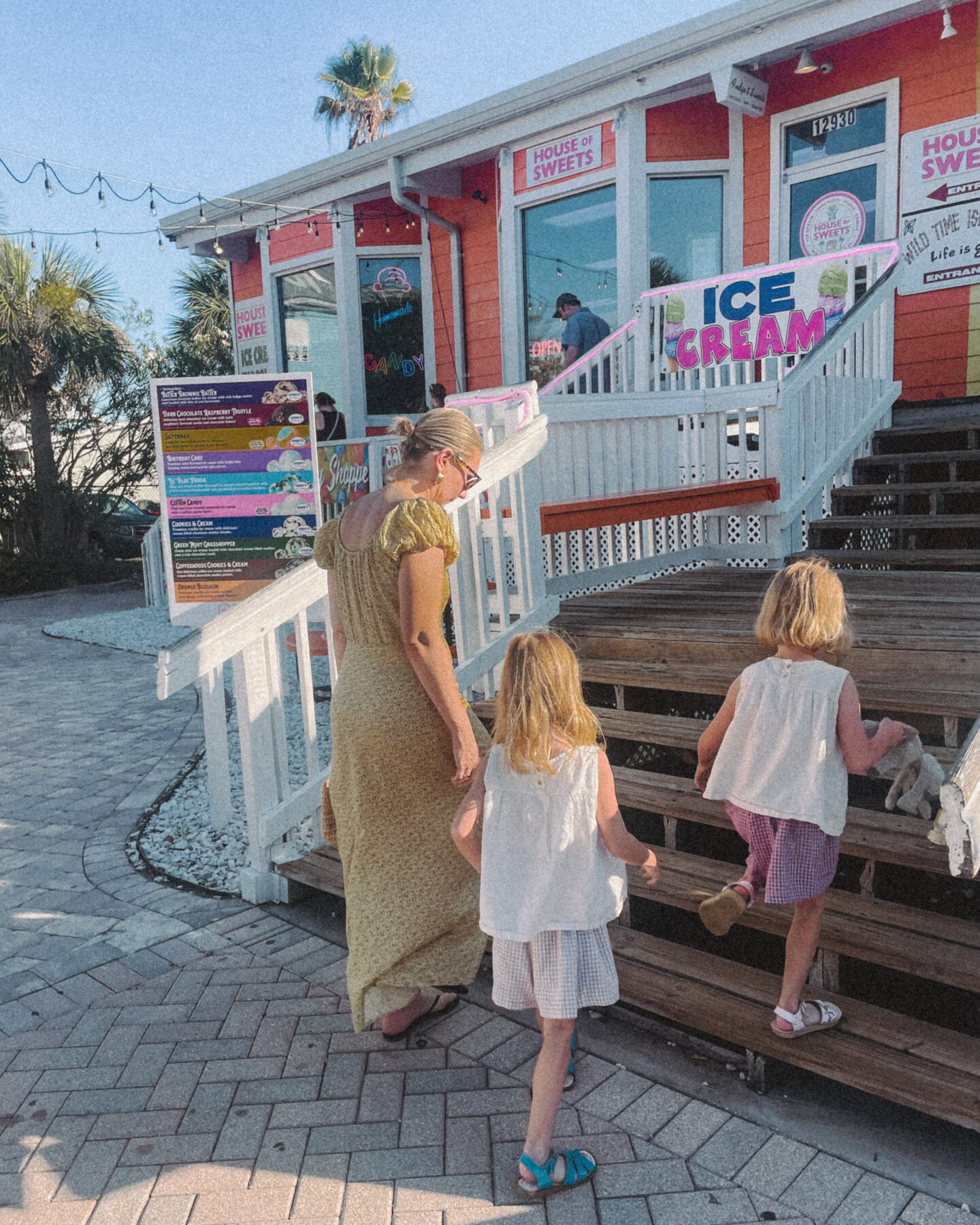 Karin Emily wearing a long yellow dress from Doen walks into a beach candy shop with her twin daughters