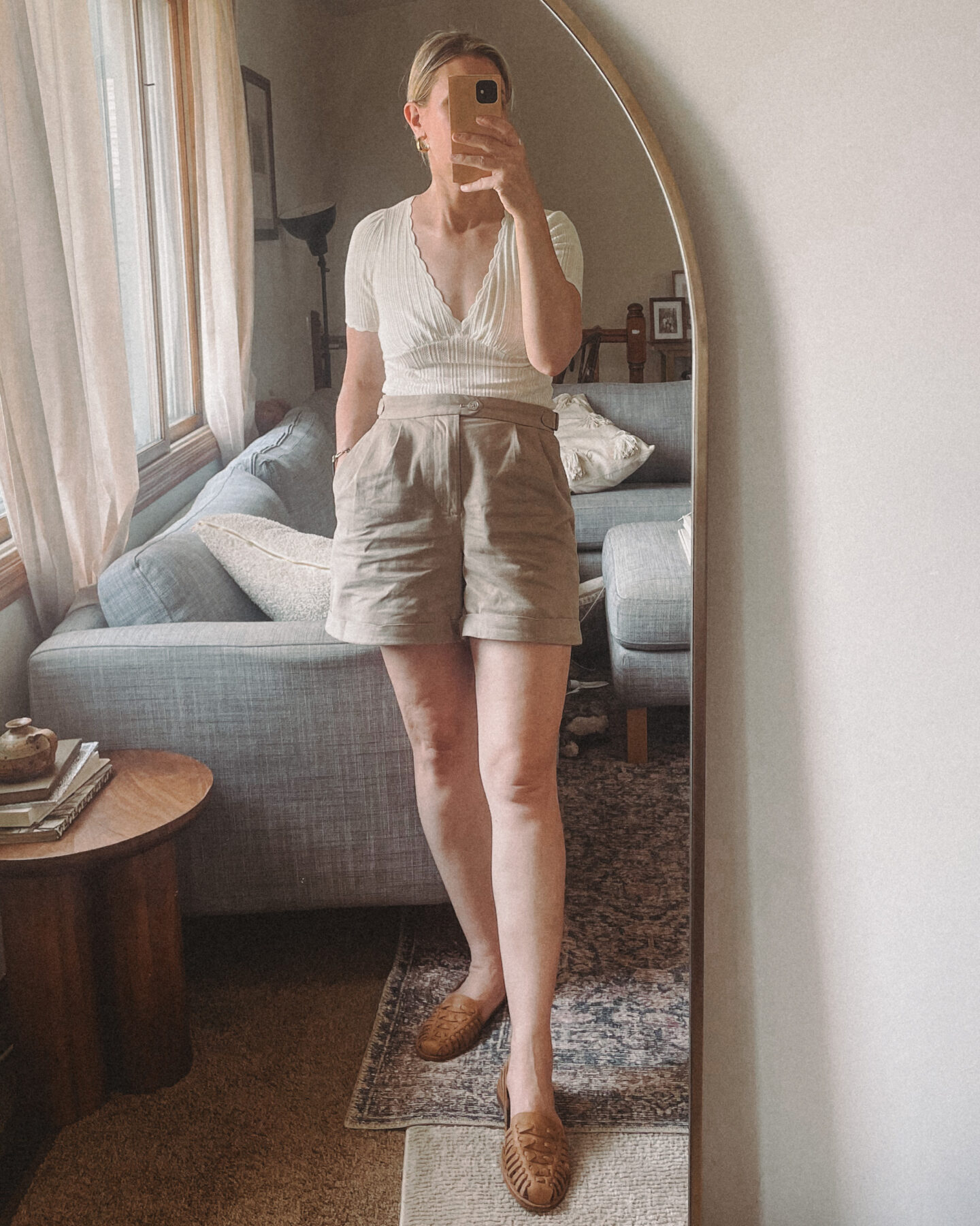 Karin Emily takes a mirror selfie in her living room A Sezane tee, pleated shorts , and Nisolo sandals 