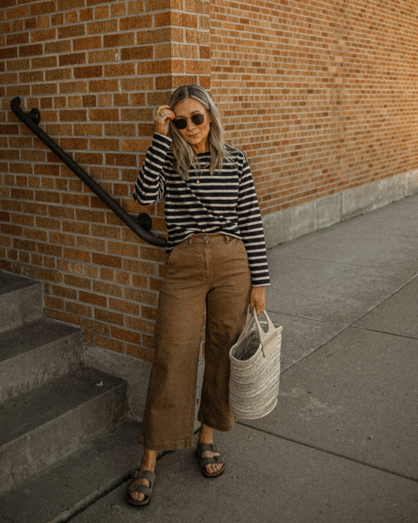 Karin. Emily stands in front of a brick wall, and where is a striped long sleeve tea from Everlane and a pair of Brown wide leg pants, and Arizona sandals from Birkenstock