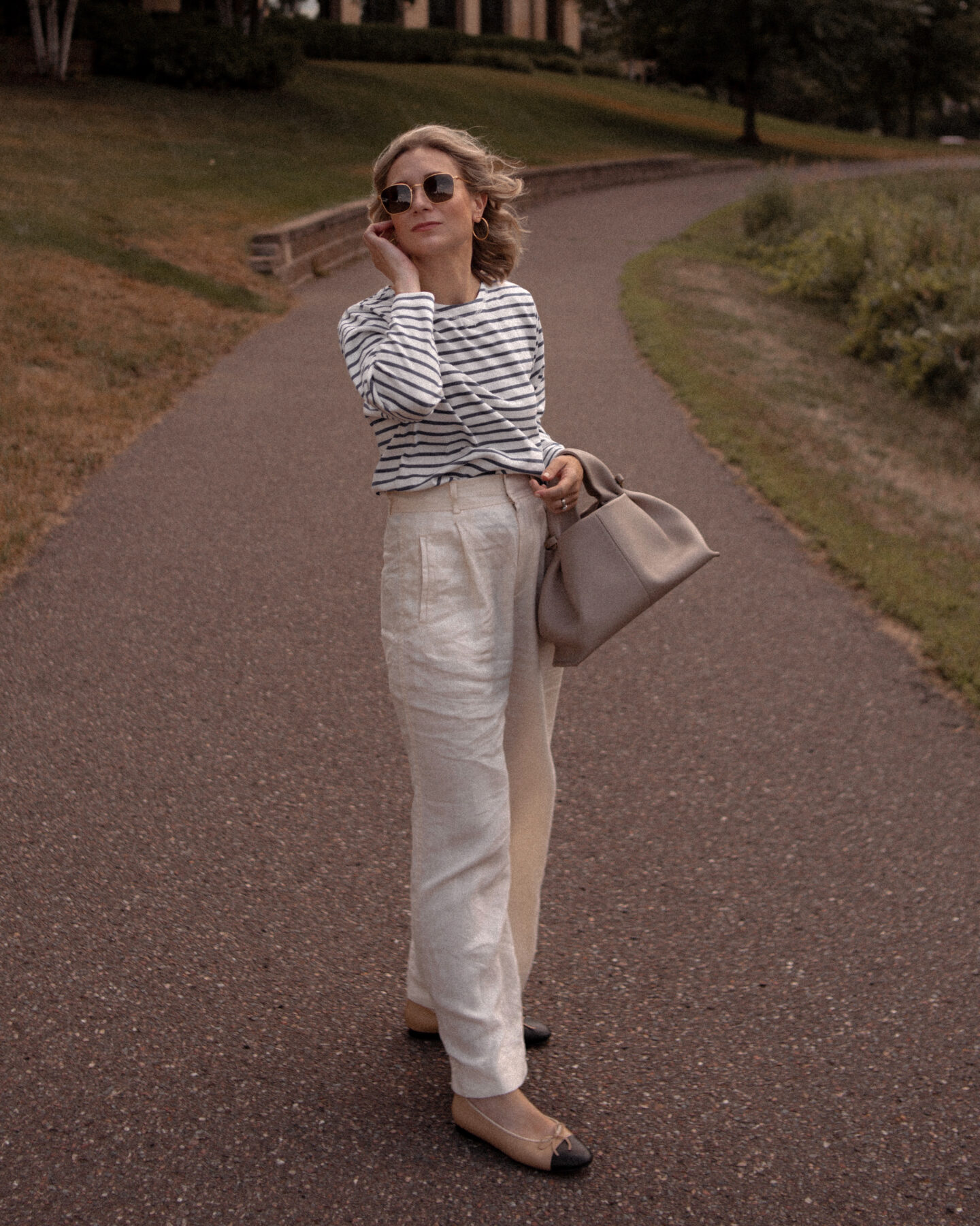 Karin Emily stands in a path and wears a blue and white striped long sleeve tee, cream wide leg linen pants and a Poline Paris bag