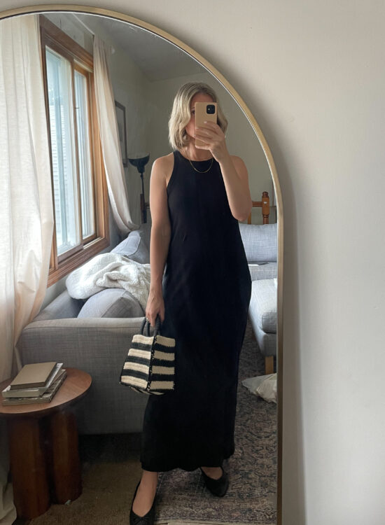 Karin Emily takes a mirror selfie wearing a black tank dress, black woven pointed toe flats and a black and natural colored straw bag