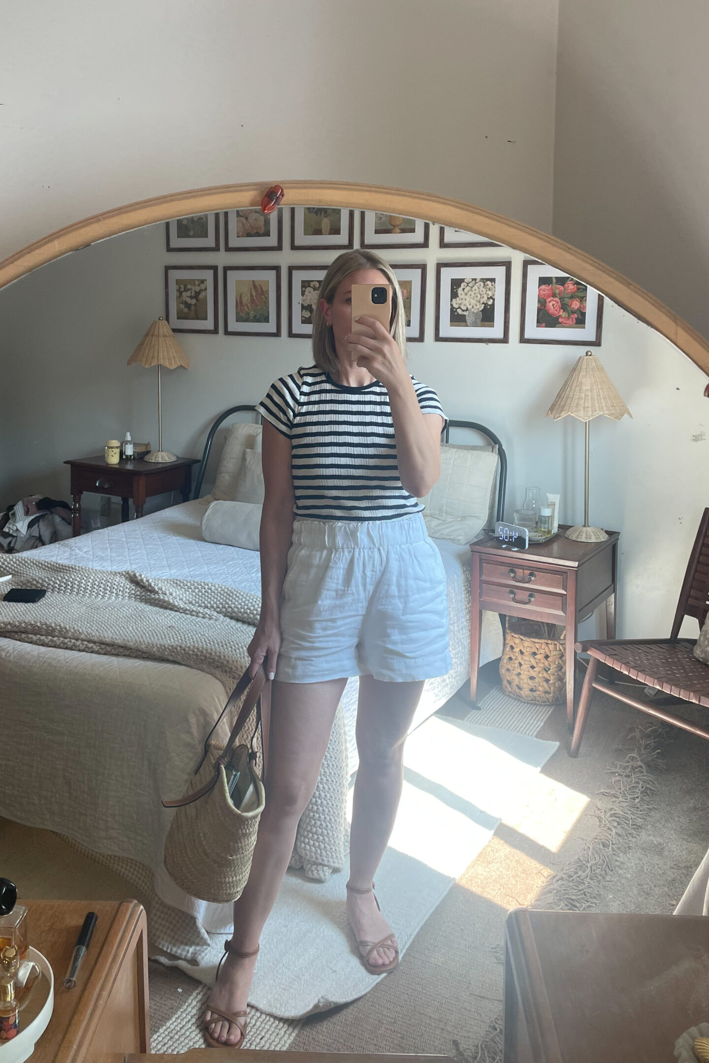 Karin Emily takes a mirror selfie wearing a striped tee from Doen, white linen pull on shorts, brown strappy sandals, and a brown basket bag