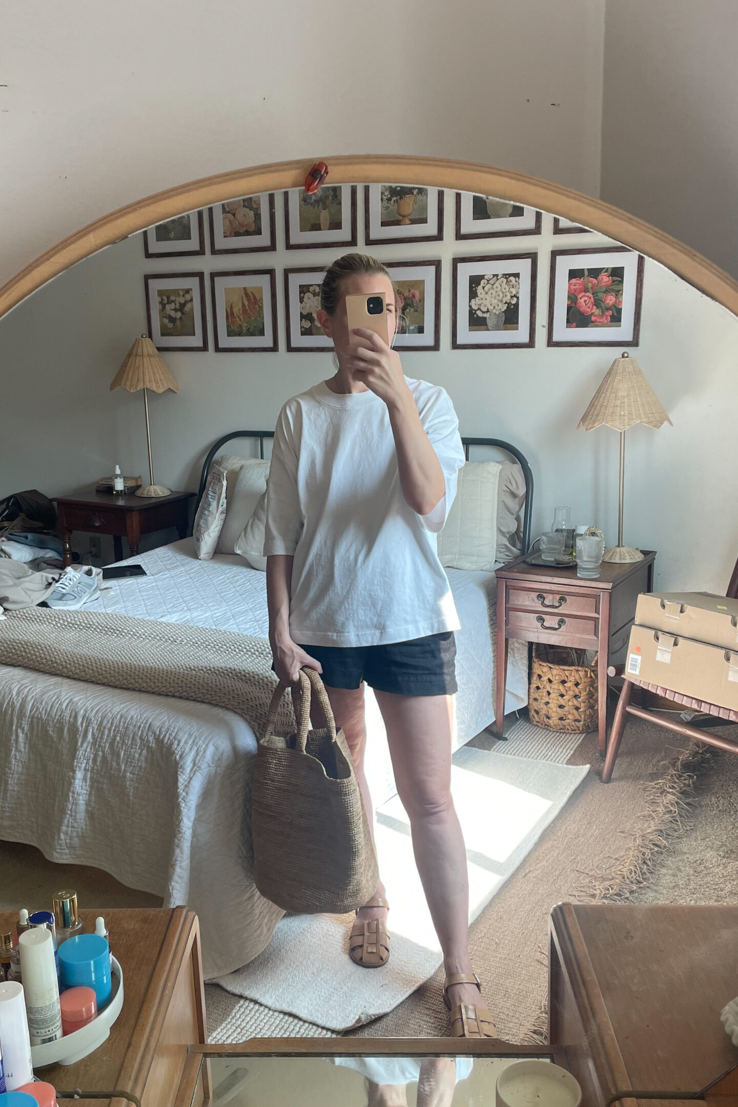 Karin Emily takes a mirror selfie in her bedroom in front of a round mirror vanity while wearing a cable knit sweater, white linen pull on shorts, and fisherman sandals from her warm weather capsule wardrobe