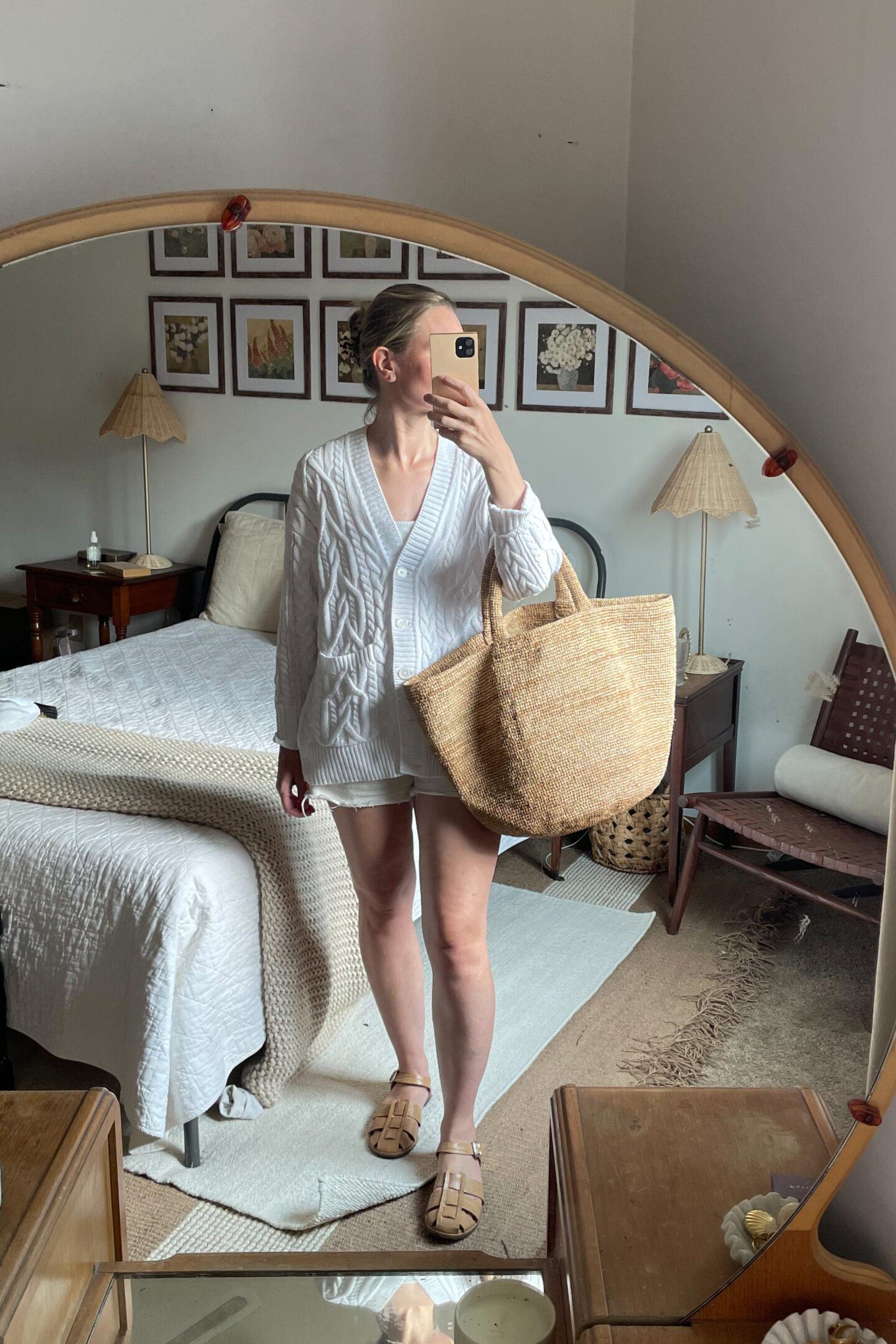 Karin Emily takes a mirror selfie wearing an oversized white cable-knit cardigan , white jean shorts, nude fisherman sandals, and a straw tote bag