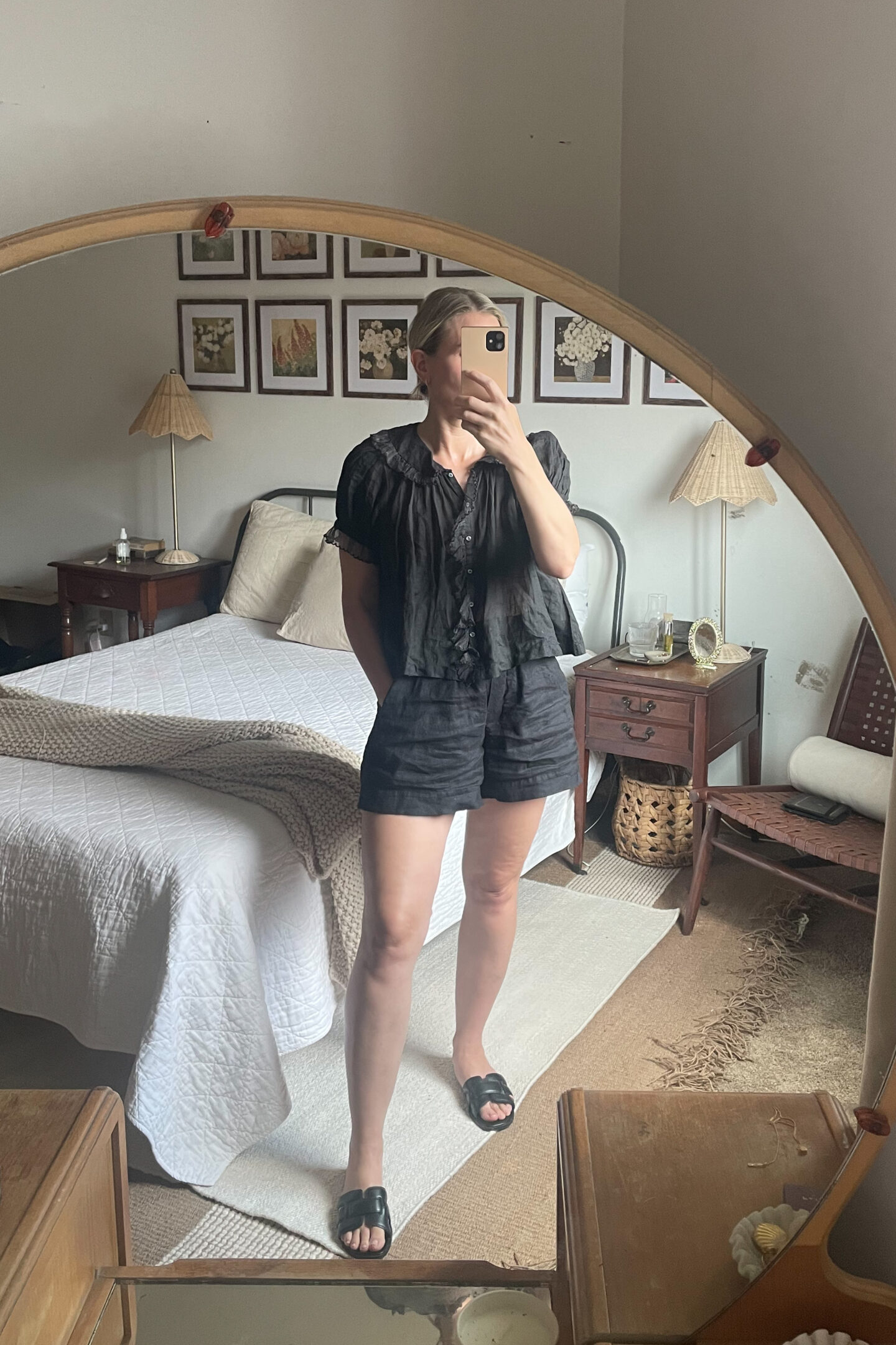 Karin Emily takes a mirror selfie and wears a black ruffled Doen top and black linen pull on shorts with black slide sandals that are YSL tribute sandal look alikes