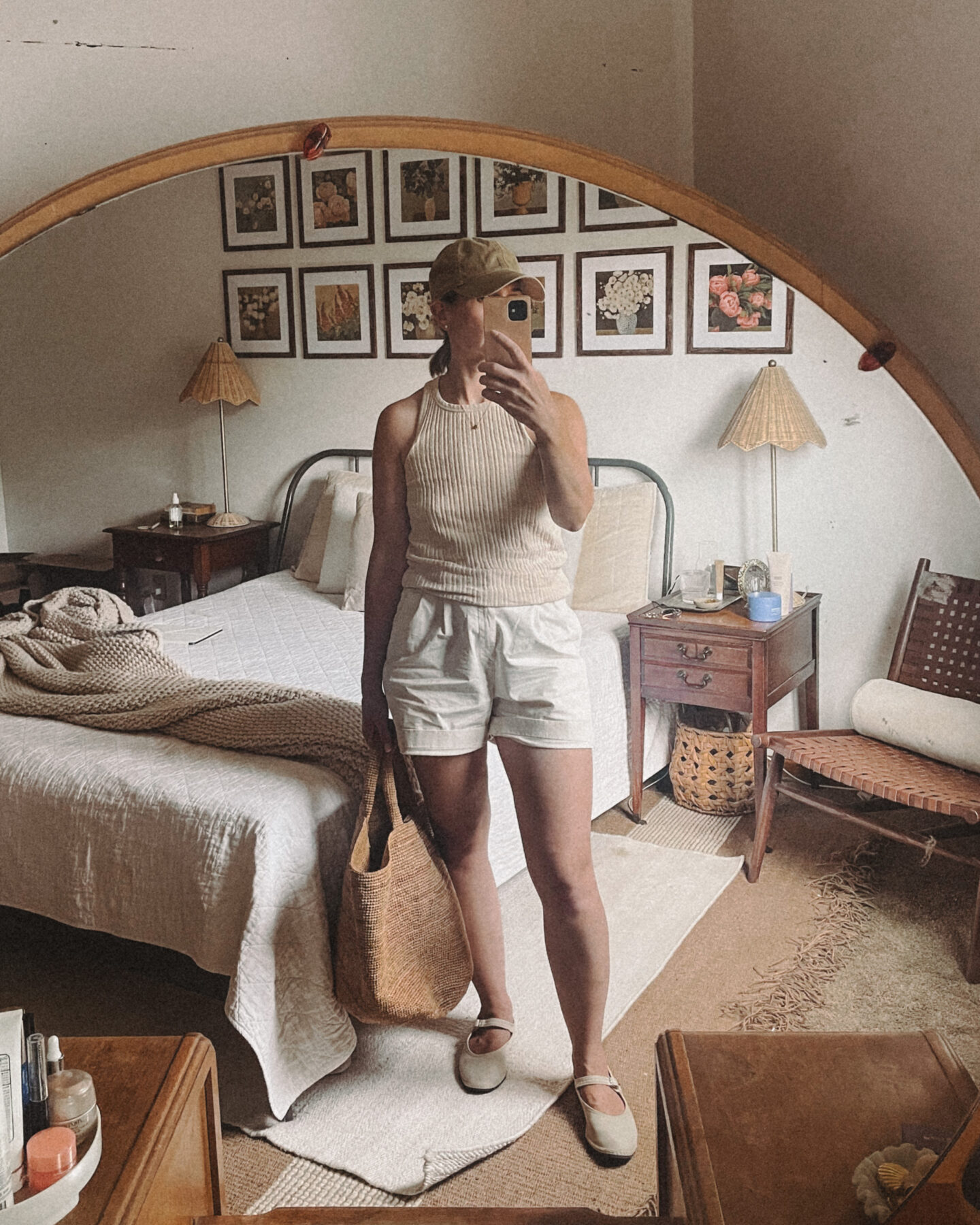 Karin Emily takes a mirror selfie in her bedroom in front of a round mirror vanity while wearing a cable knit sweater, white linen pull on shorts, and fisherman sandals from her warm weather capsule wardrobe