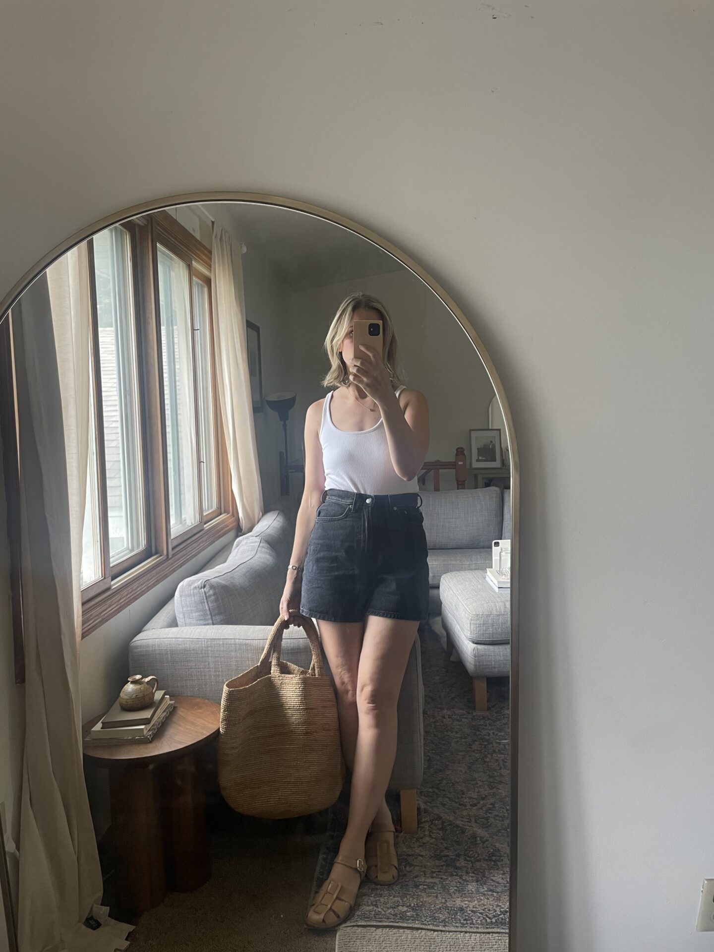 Karin Emily takes a mirror selfie in her bedroom in front of a round mirror vanity while wearing a cable knit sweater, white linen pull on shorts, and fisherman sandals from her warm weather capsule wardrobe