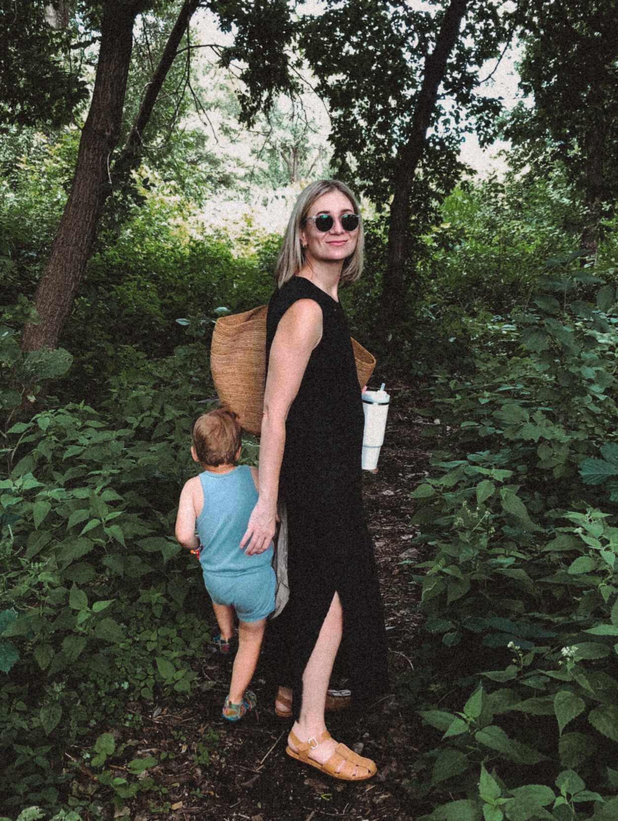 Karin Emily stands in the woods in a black dress from her capsule wardrobe with her son who’s in a blue romper