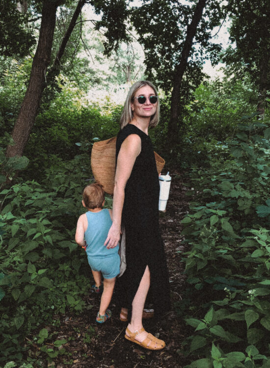 Karin Emily stands in the woods in a black dress from her capsule wardrobe with her son who’s in a blue romper