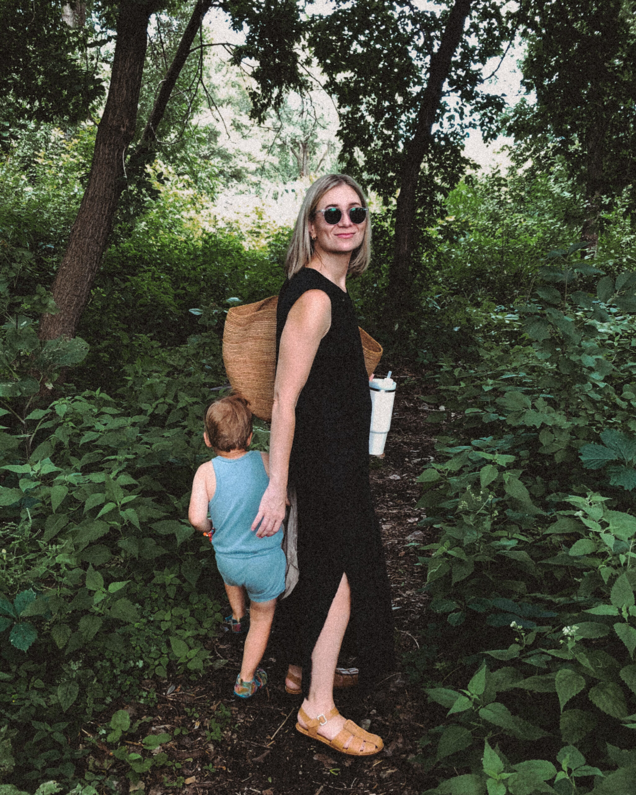 Karin Emily stands in the woods in a black dress from her capsule wardrobe with her son who’s in a blue romper