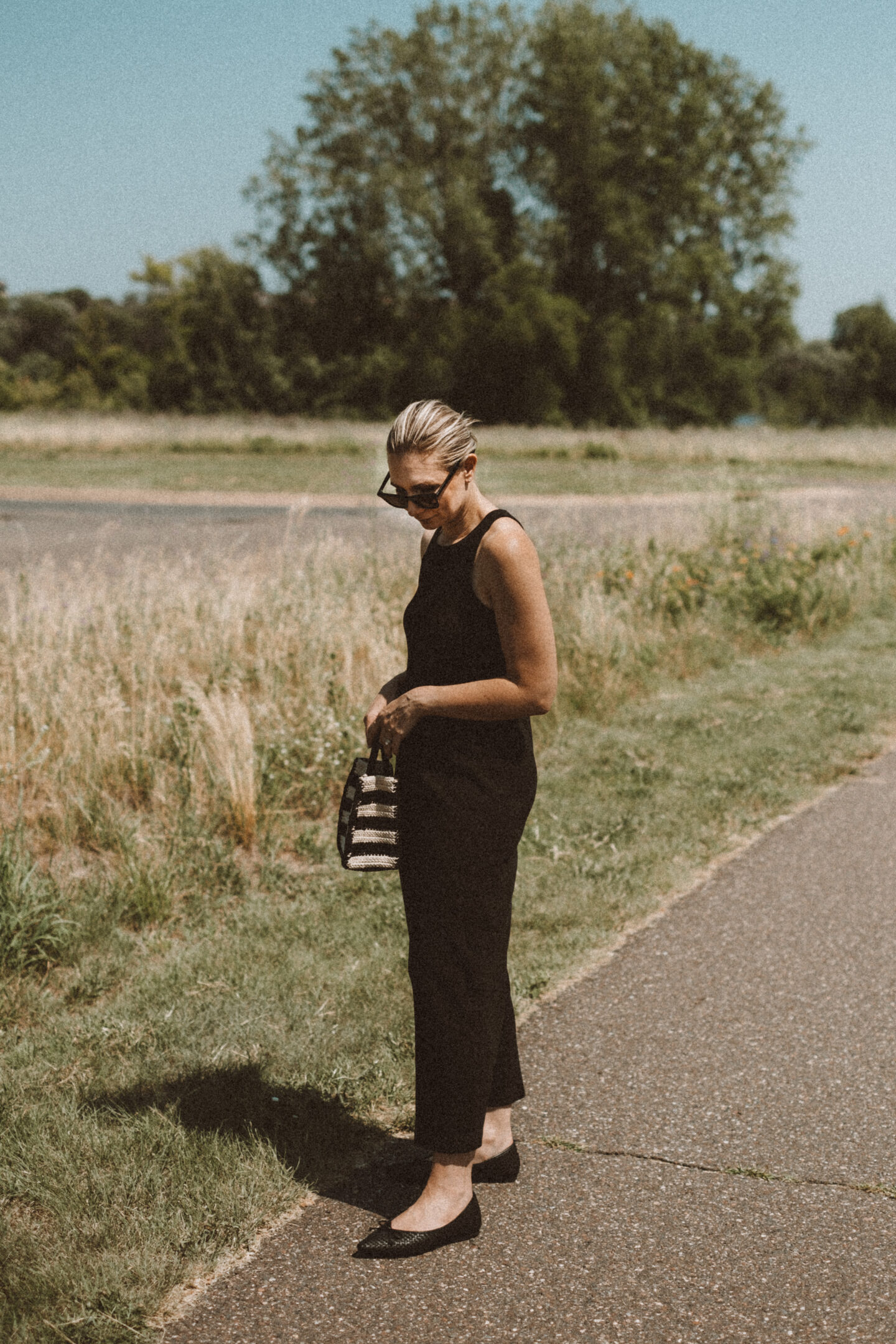 Karin Emily wears a black tank dress and the Birdies Goldfinch flats while standing on a sidewalk in front of a field of wild grasses 