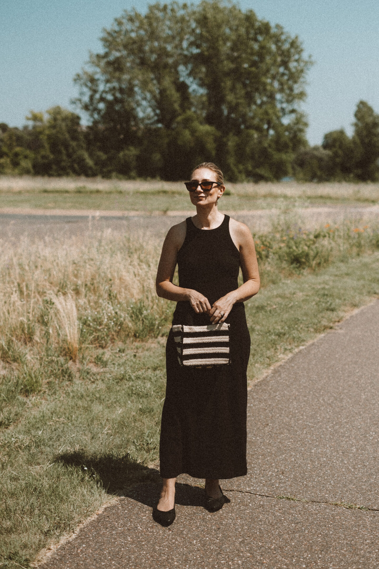 Karin Emily wears a black tank dress and the Birdies Goldfinch flats while standing on a sidewalk in front of a field of wild grasses 