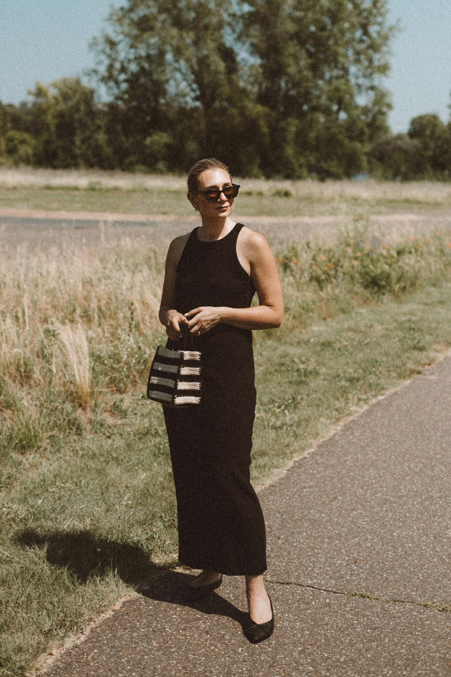 Karin Emily wears a black tank dress and the Birdies Goldfinch flats while standing on a sidewalk in front of a field of wild grasses 