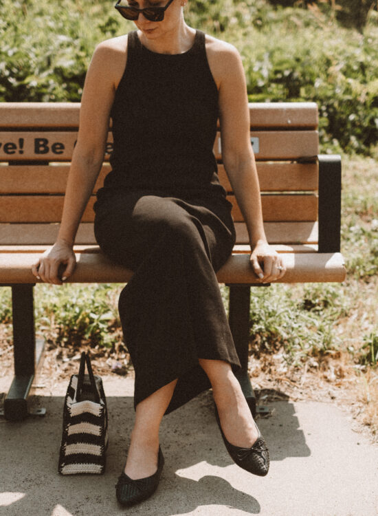 Karin Emily wears a black tank dress and the Birdies Goldfinch flats while standing on a sidewalk in front of a field of wild grasses