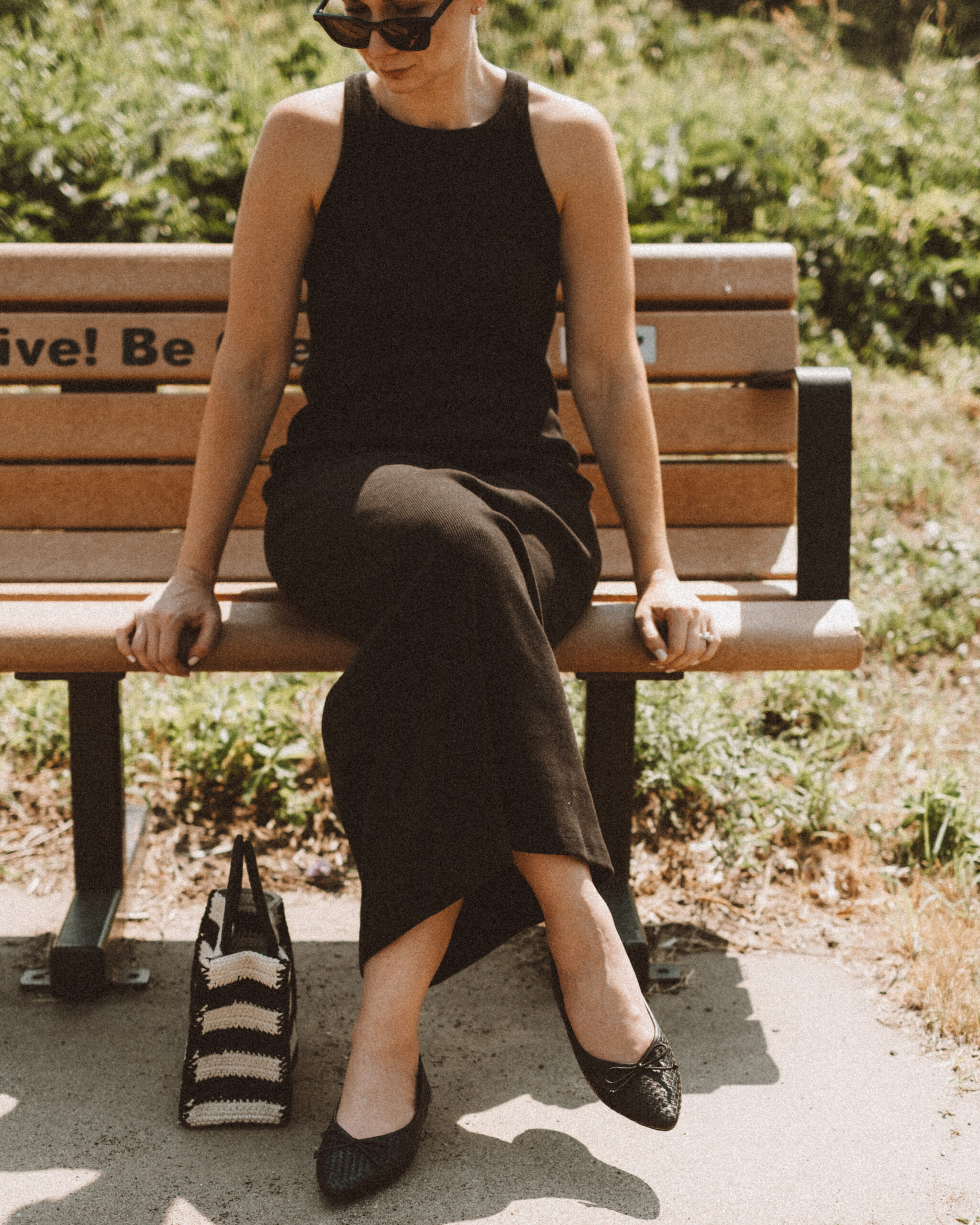 Karin Emily wears a black tank dress and the Birdies Goldfinch flats while standing on a sidewalk in front of a field of wild grasses