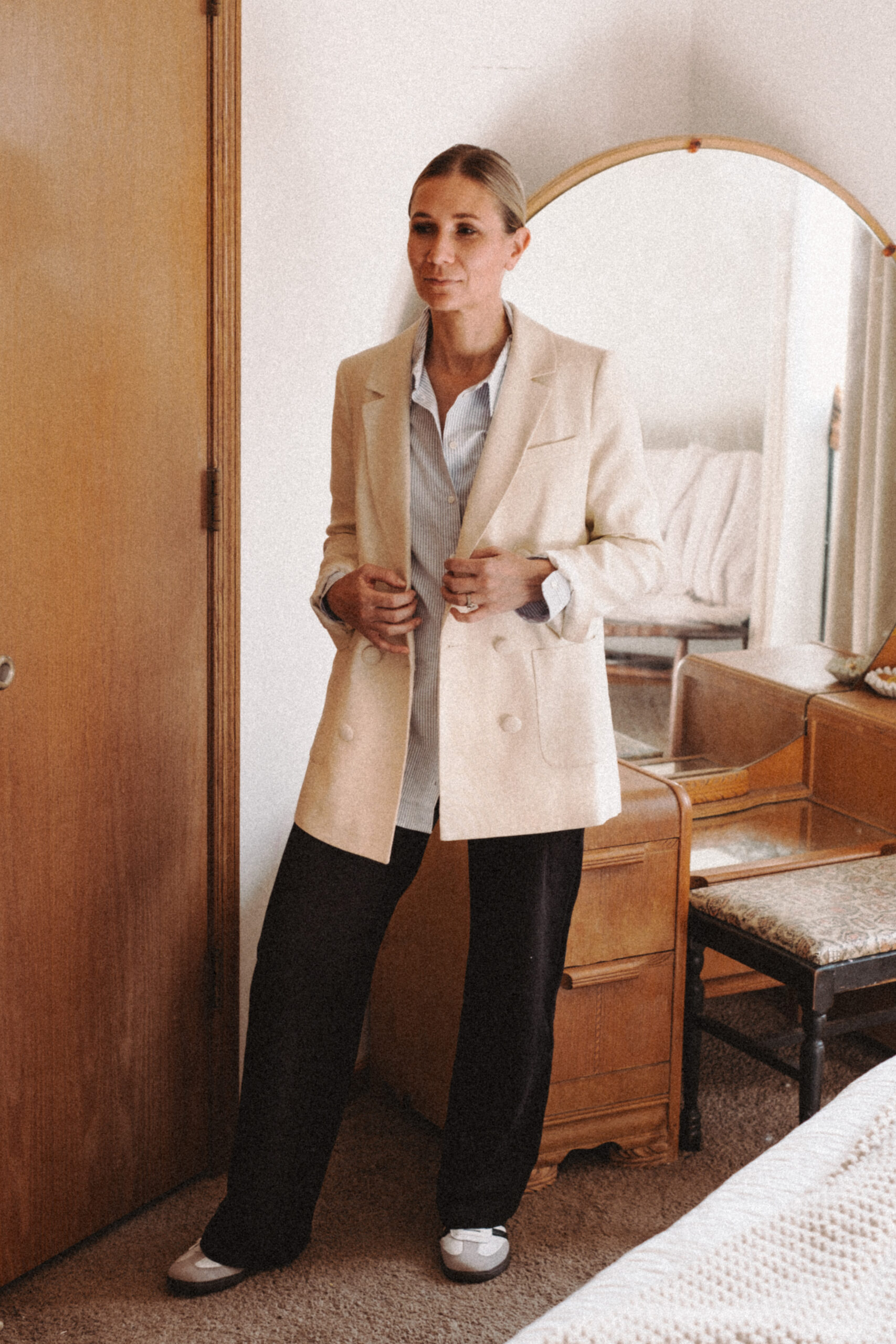 Karin Emily stands in front of her mid century vanity wearing an oversized blazer, a blue striped button down shirt, black wide leg pants, and adidas samba sneakers 