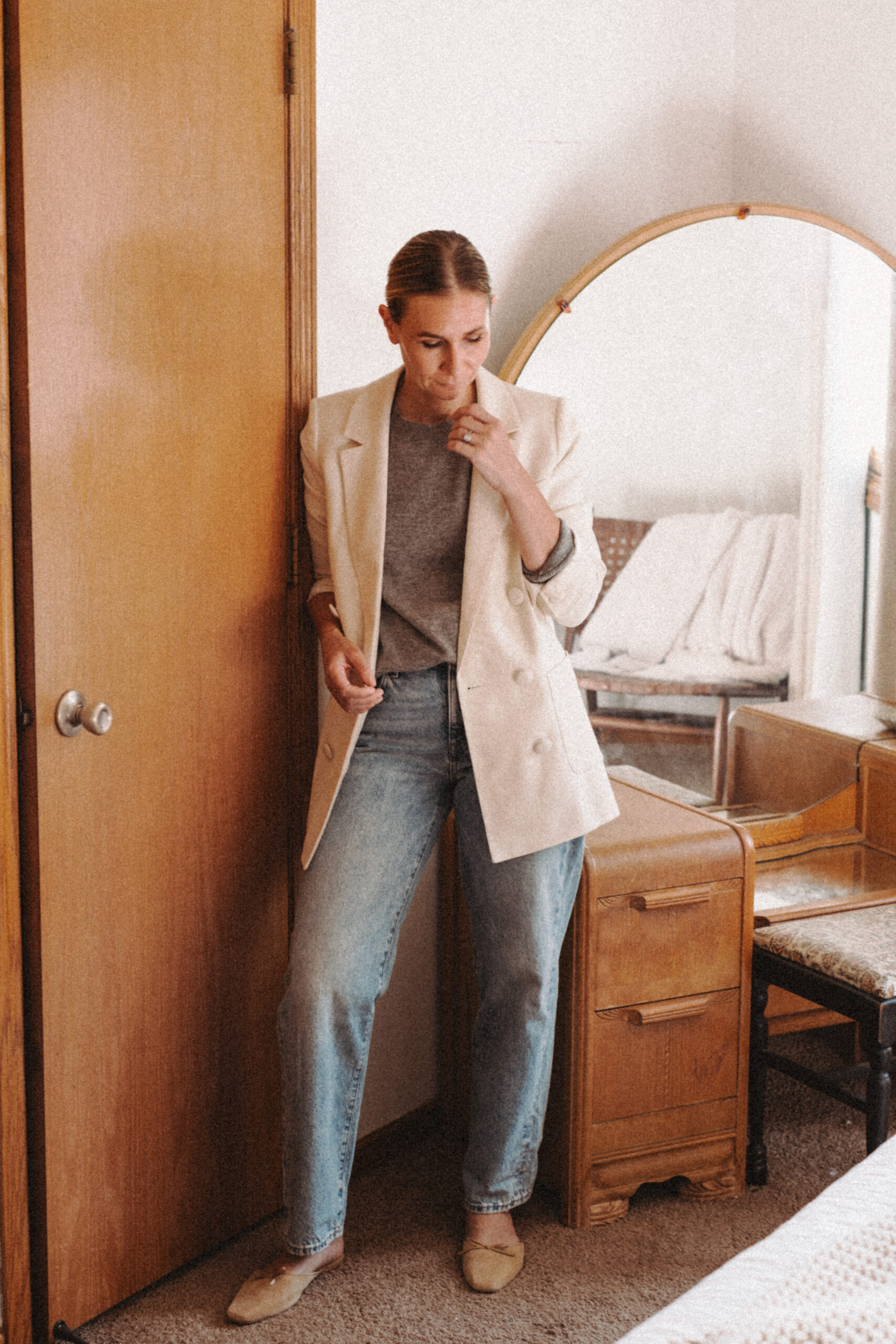 Karin Emily stands in front of her mid century vanity wearing an oversized blazer, a gray cashmere sweater, relaxed jeans from Madewell and mule flats from Doen