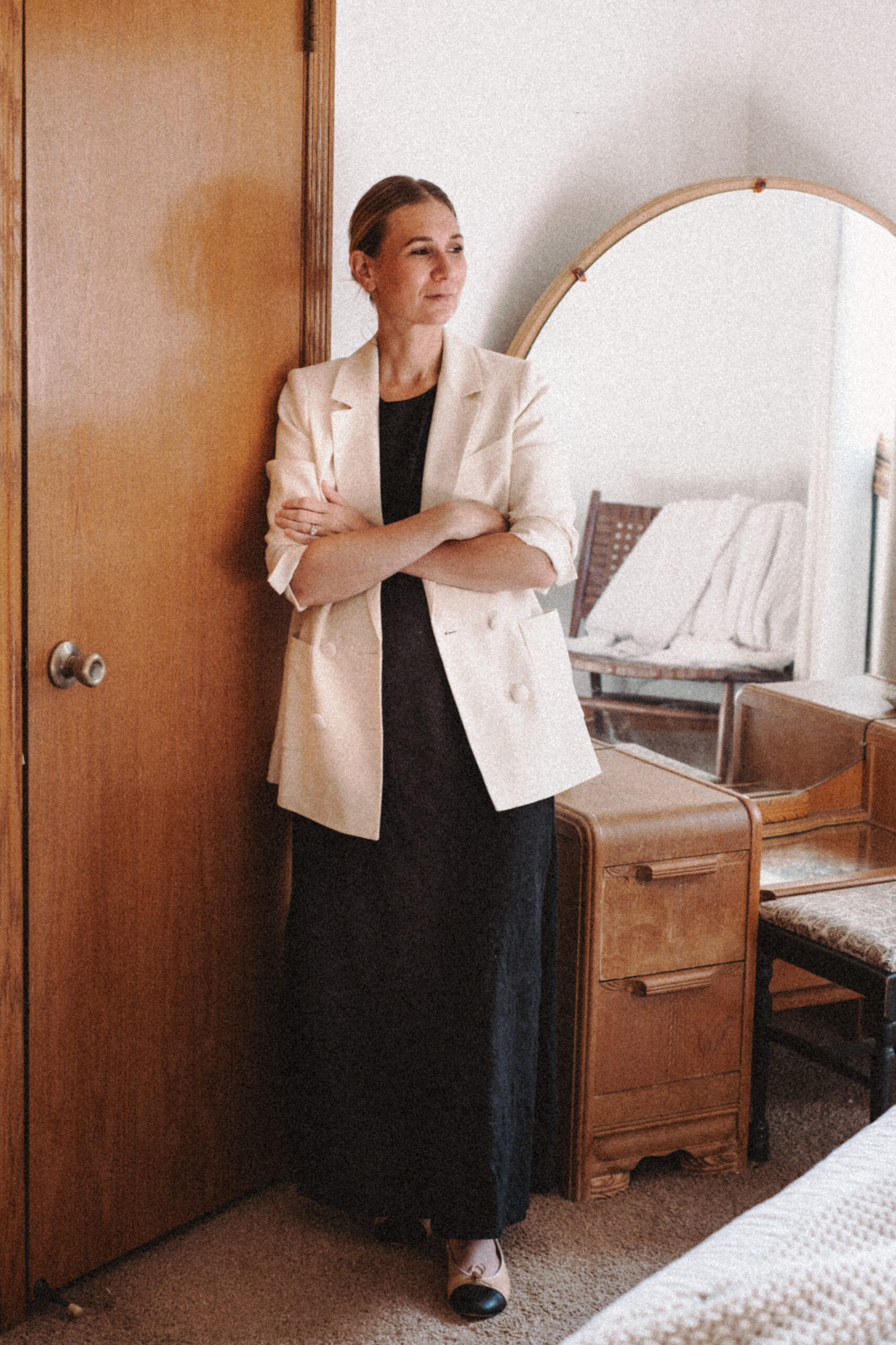 Karin Emily stands in front of her mid century vanity wearing an oversized blazer With a little black dress and Chanel look alike flats