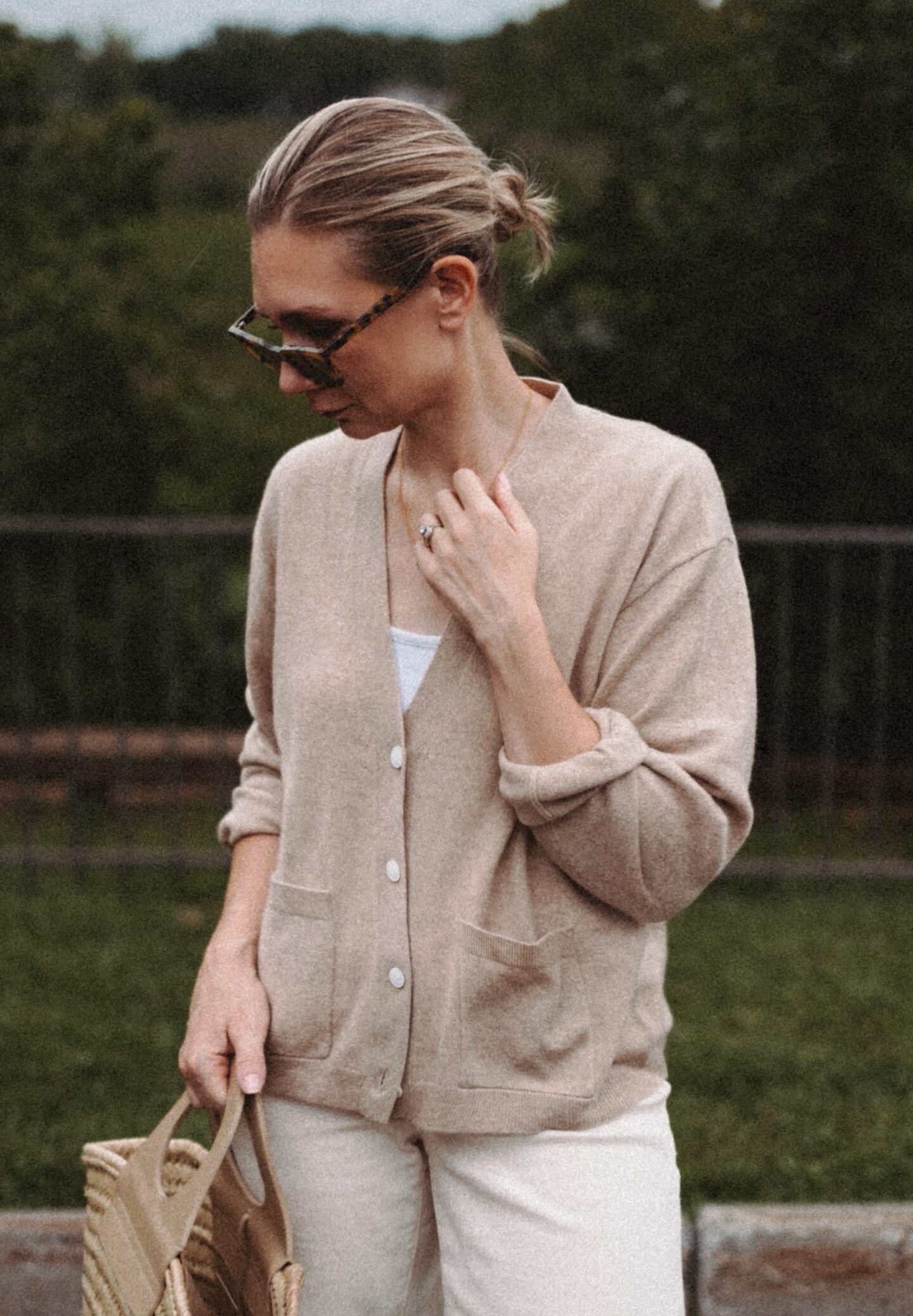 Karin Emily wears a cashmere cardigan with a pair of wide leg jeans and a Hereu basket bag in front of a wooded valley and fence