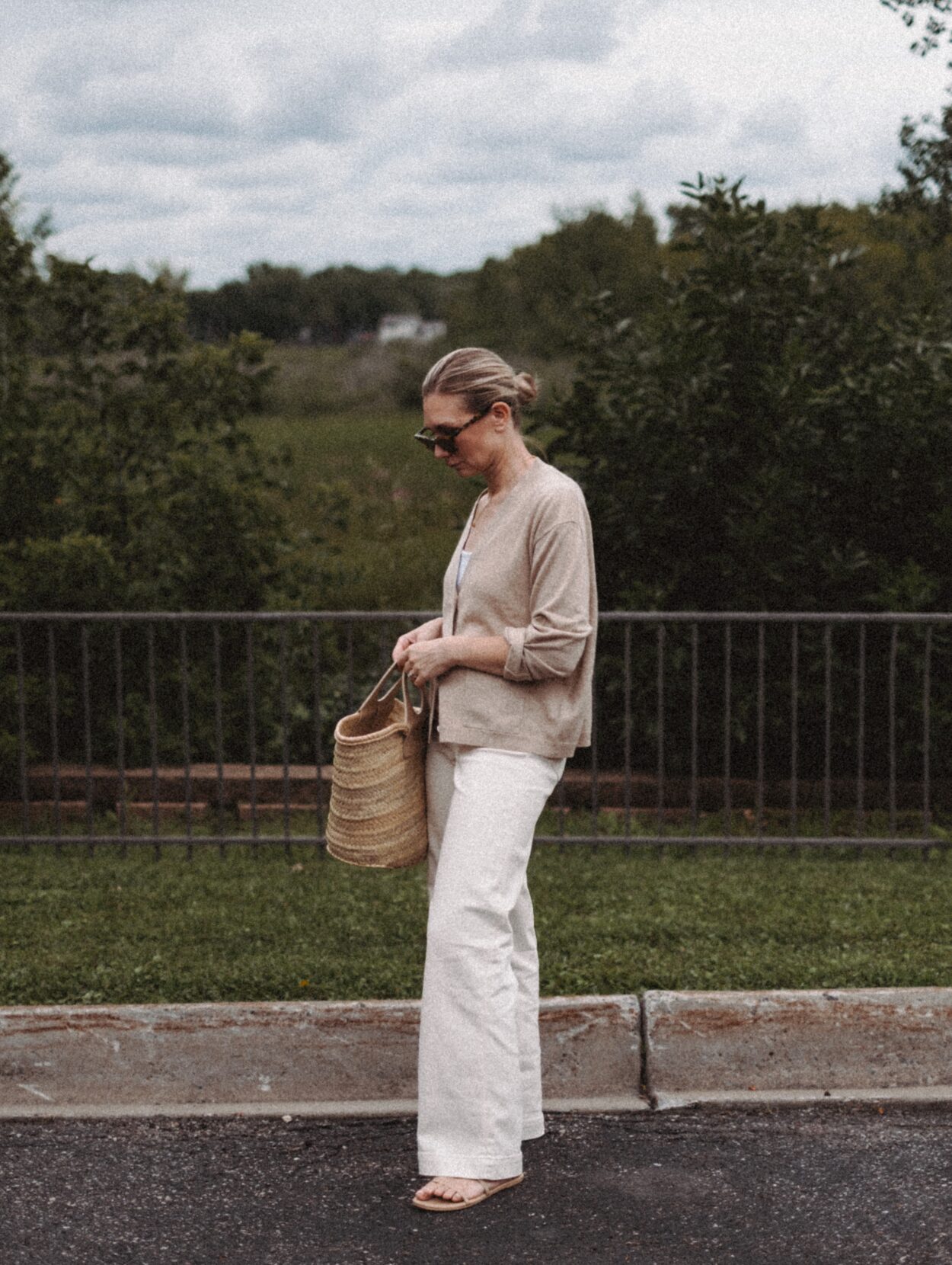 Karin Emily wears a cashmere cardigan with a pair of wide leg jeans and a Hereu basket bag in front of a wooded valley and fence