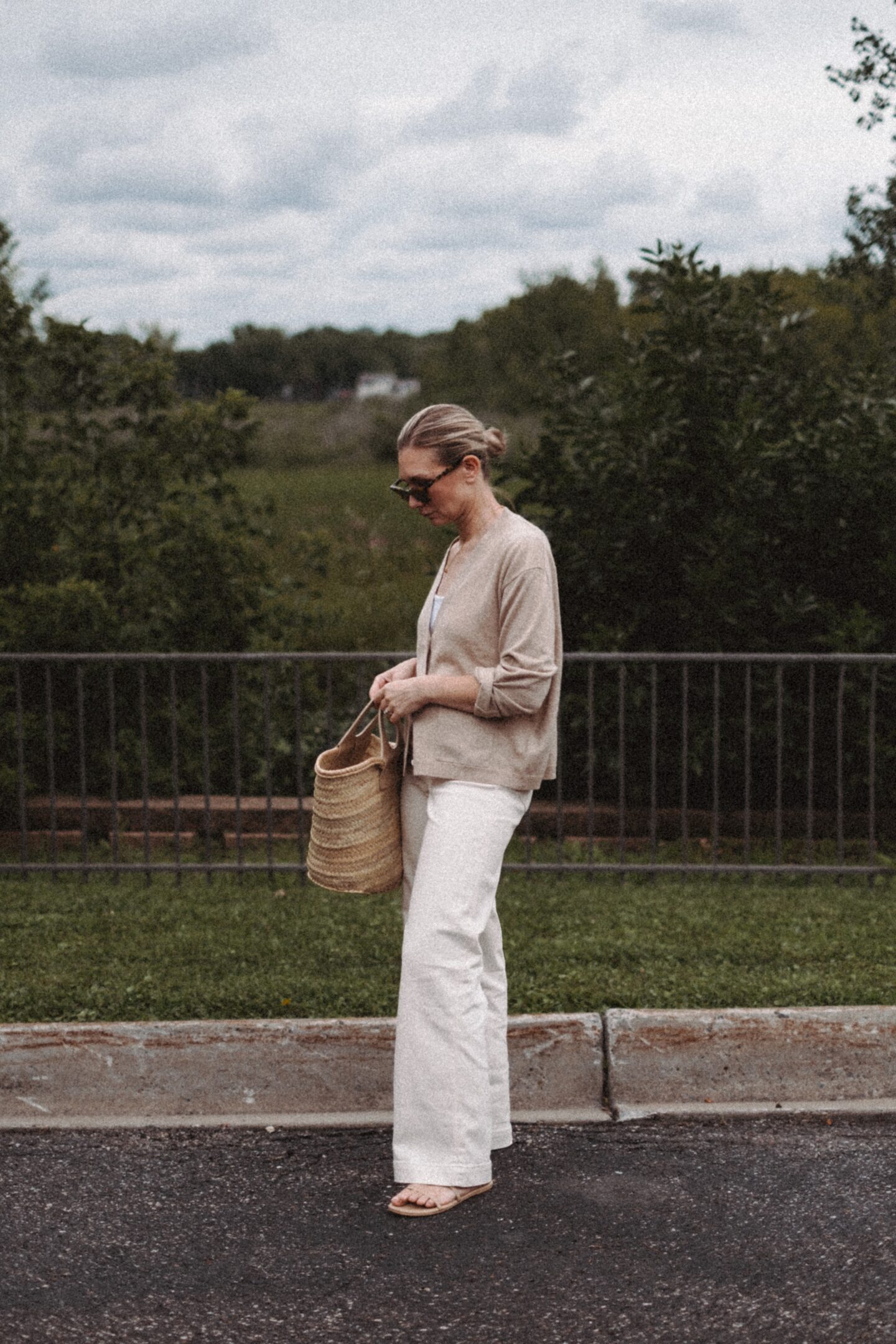 Karin Emily wears a cashmere cardigan with a pair of wide leg jeans and a Hereu basket bag in front of a wooded valley and fence