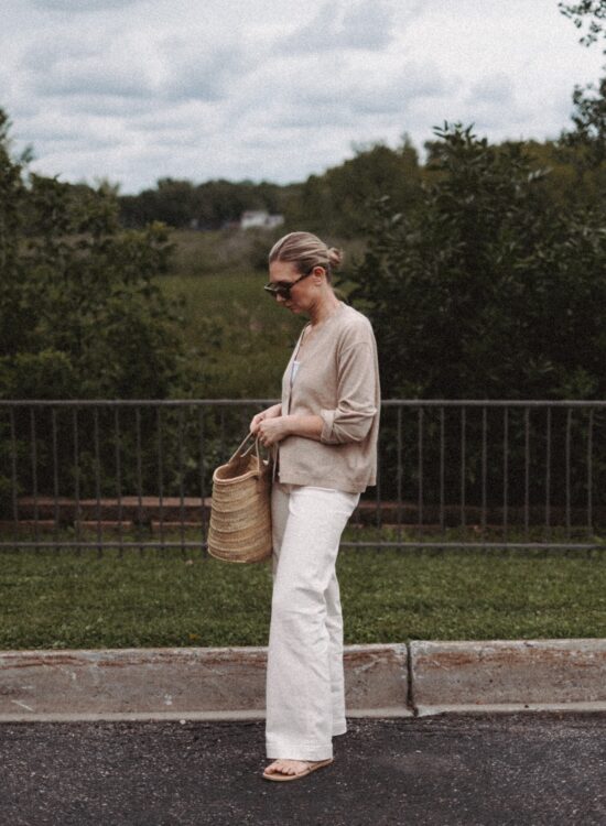 Karin Emily wears a cashmere cardigan with a pair of wide leg jeans and a Hereu basket bag in front of a wooded valley and fence