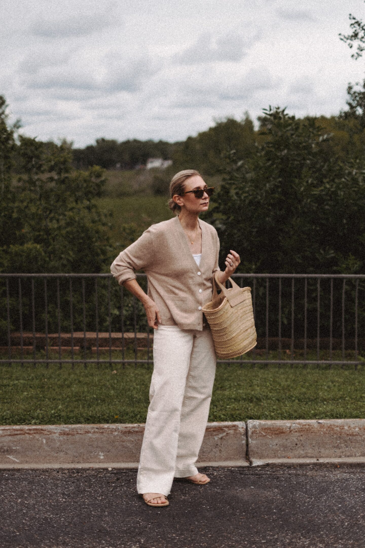 Karin Emily wears a cashmere cardigan with a pair of wide leg jeans and a Hereu basket bag in front of a wooded valley and fence