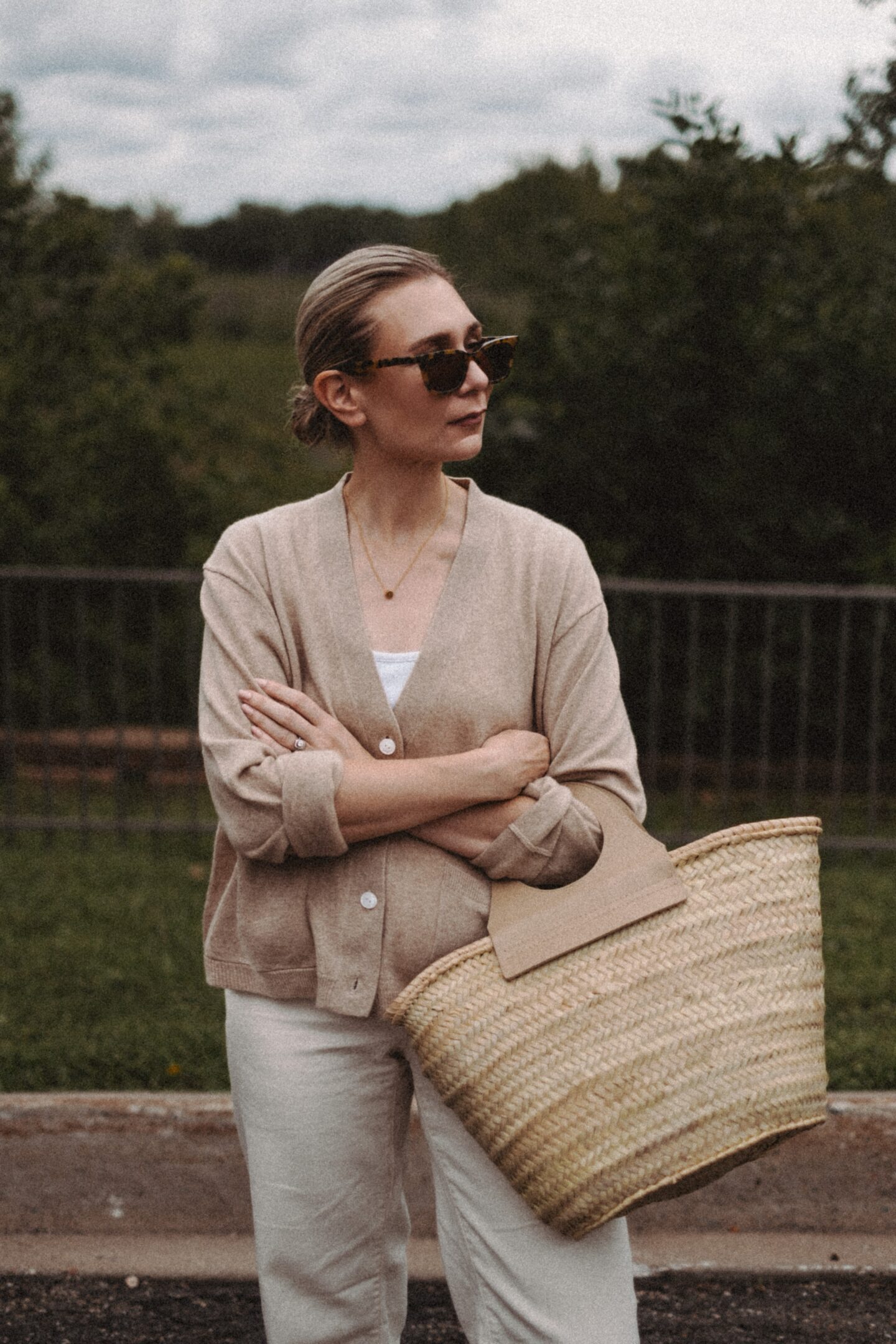 Karin Emily wears a cashmere cardigan with a pair of wide leg jeans and a Hereu basket bag in front of a wooded valley and fence