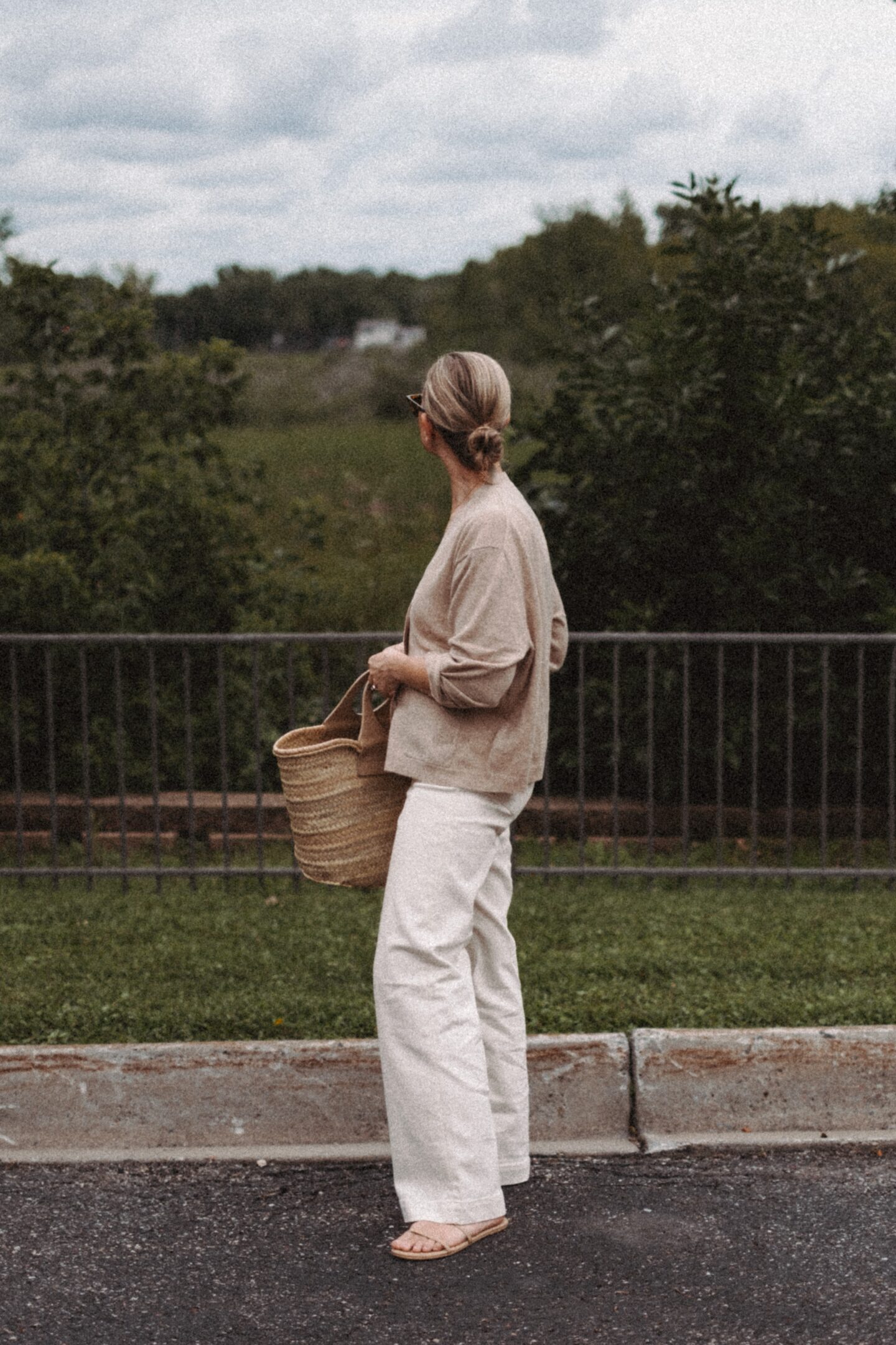 Karin Emily wears a cashmere cardigan with a pair of wide leg jeans and a Hereu basket bag in front of a wooded valley and fence