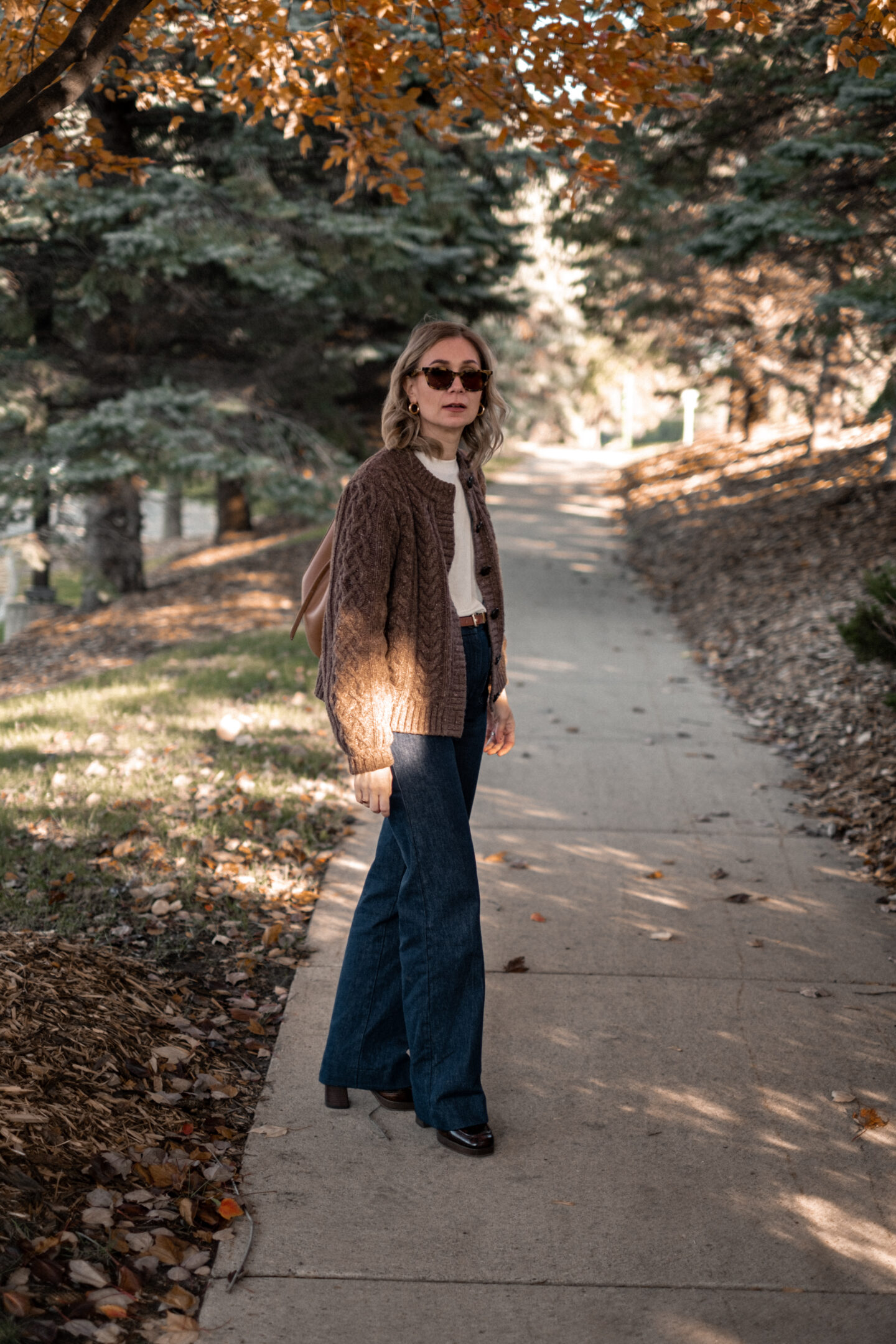 Karin Emily stands on a wooded pathway with fall colors wearing a pair of platform loafers wearing a park of dark wash trouser jeans, a chocolate brown cable knit cardigan all from Sezane, and a brown Polene bag