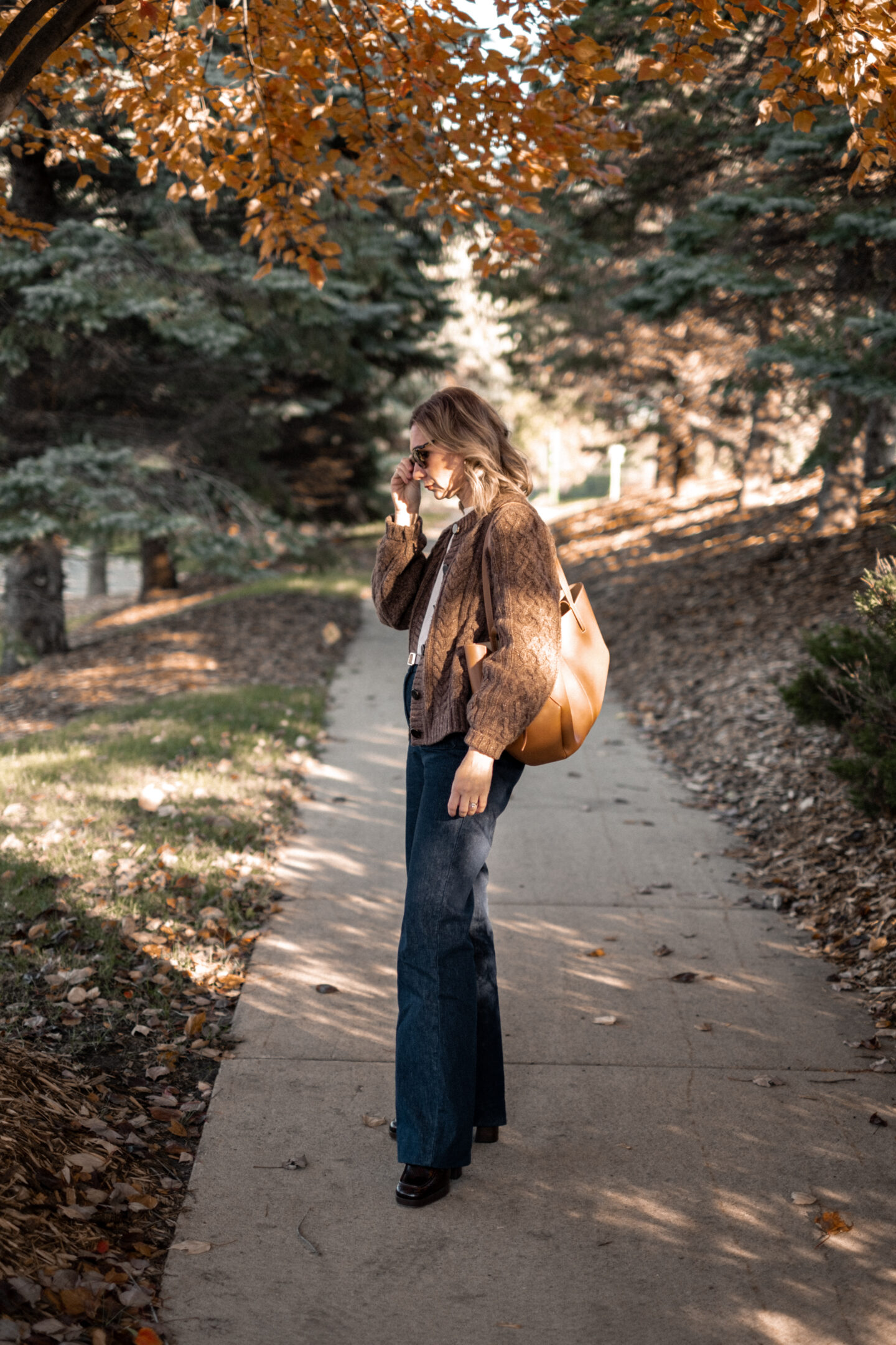 Karin Emily stands on a wooded pathway with fall colors wearing a pair of platform loafers wearing a park of dark wash trouser jeans, a chocolate brown cable knit cardigan all from Sezane, and a brown Polene bag