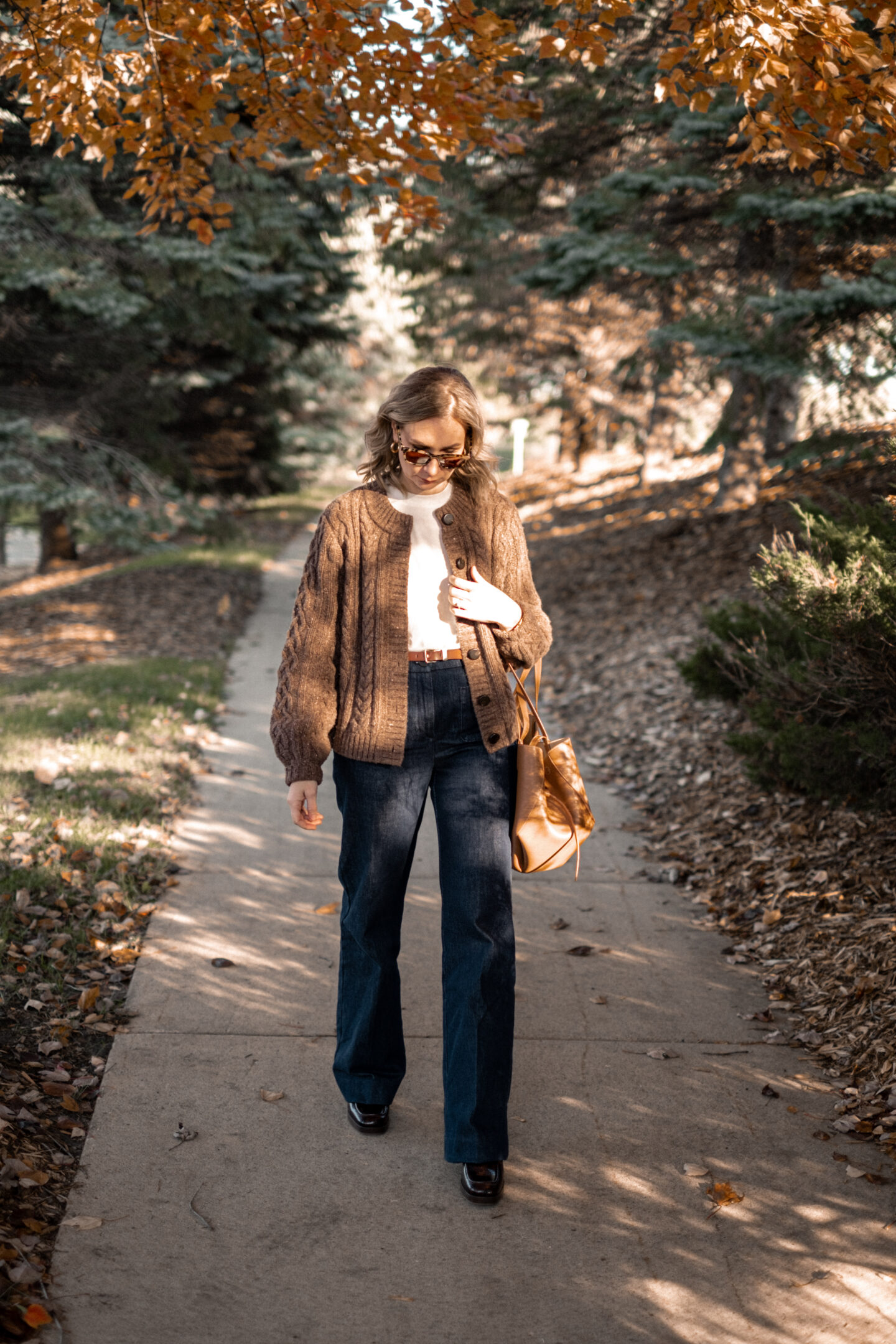 Karin Emily stands on a wooded pathway with fall colors wearing a pair of platform loafers wearing a park of dark wash trouser jeans, a chocolate brown cable knit cardigan all from Sezane, and a brown Polene bag