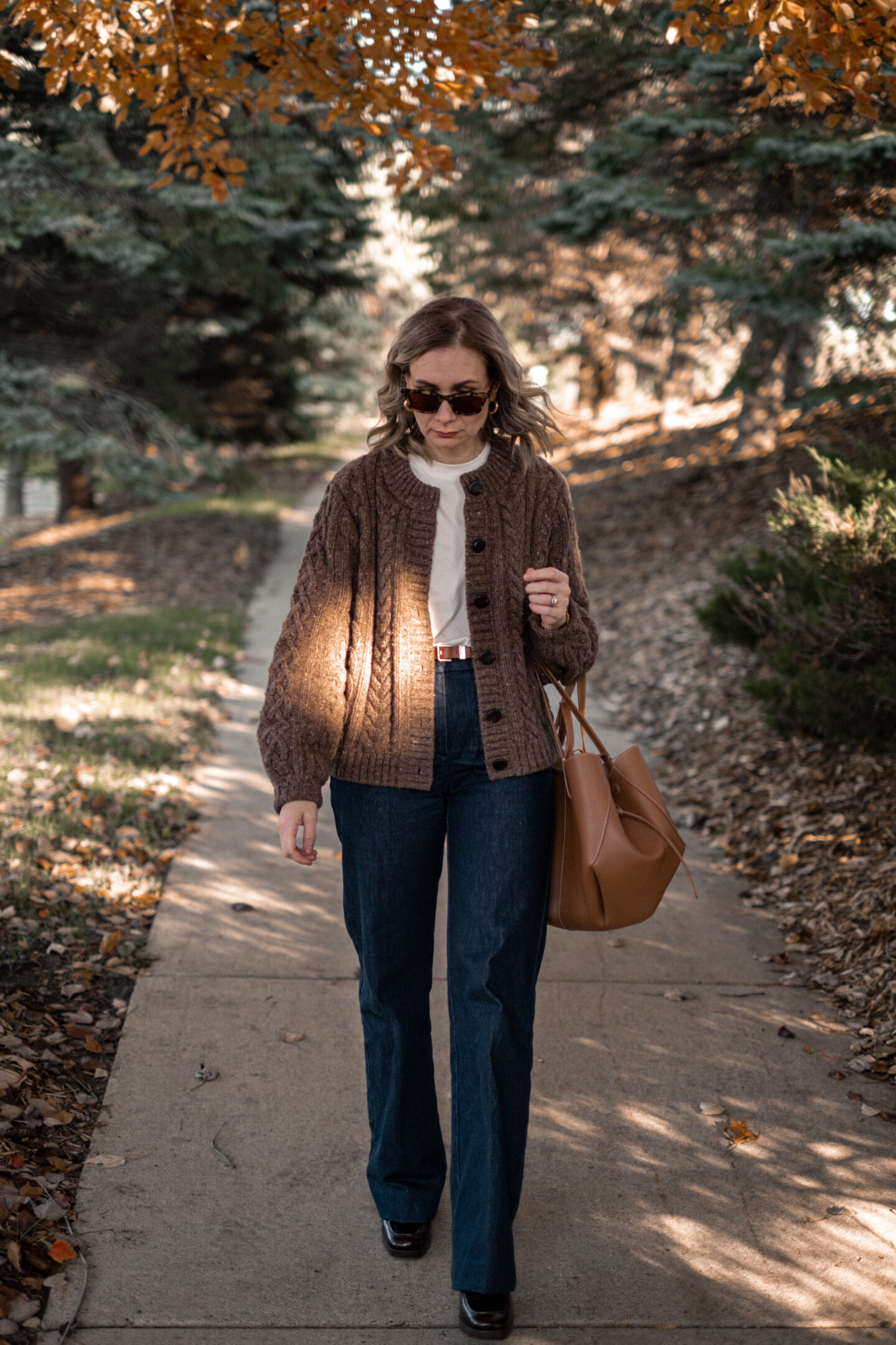 Karin Emily stands on a wooded pathway with fall colors wearing a pair of platform loafers wearing a park of dark wash trouser jeans, a chocolate brown cable knit cardigan all from Sezane, and a brown Polene bag