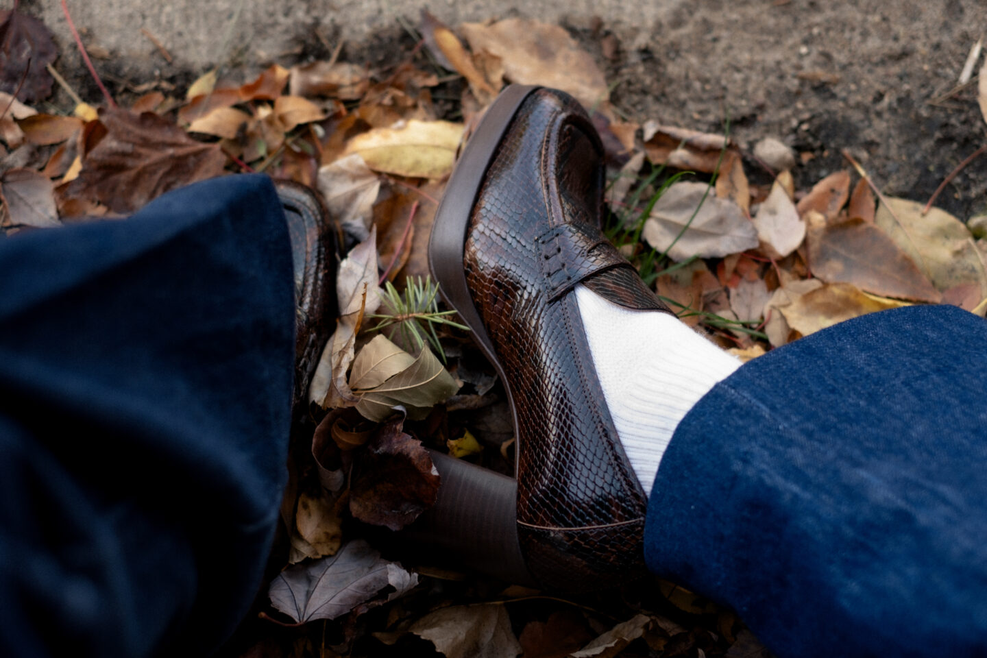 Karin Emily stands on a wooded pathway with fall colors wearing a pair of platform loafers wearing a park of dark wash trouser jeans, a chocolate brown cable knit cardigan all from Sezane, and a brown Polene bag