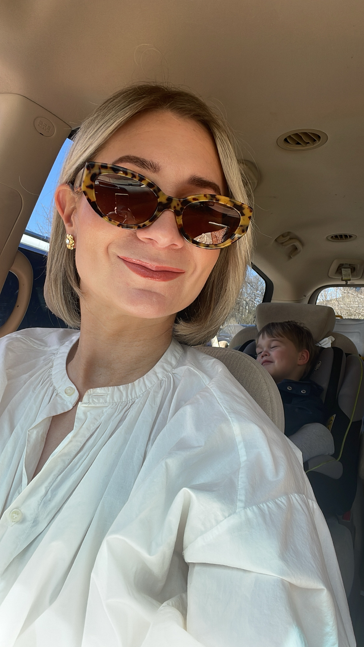 Karin Emily wears a pair of sunglasses in her car with a pair of gold flower earrings and a white blouse