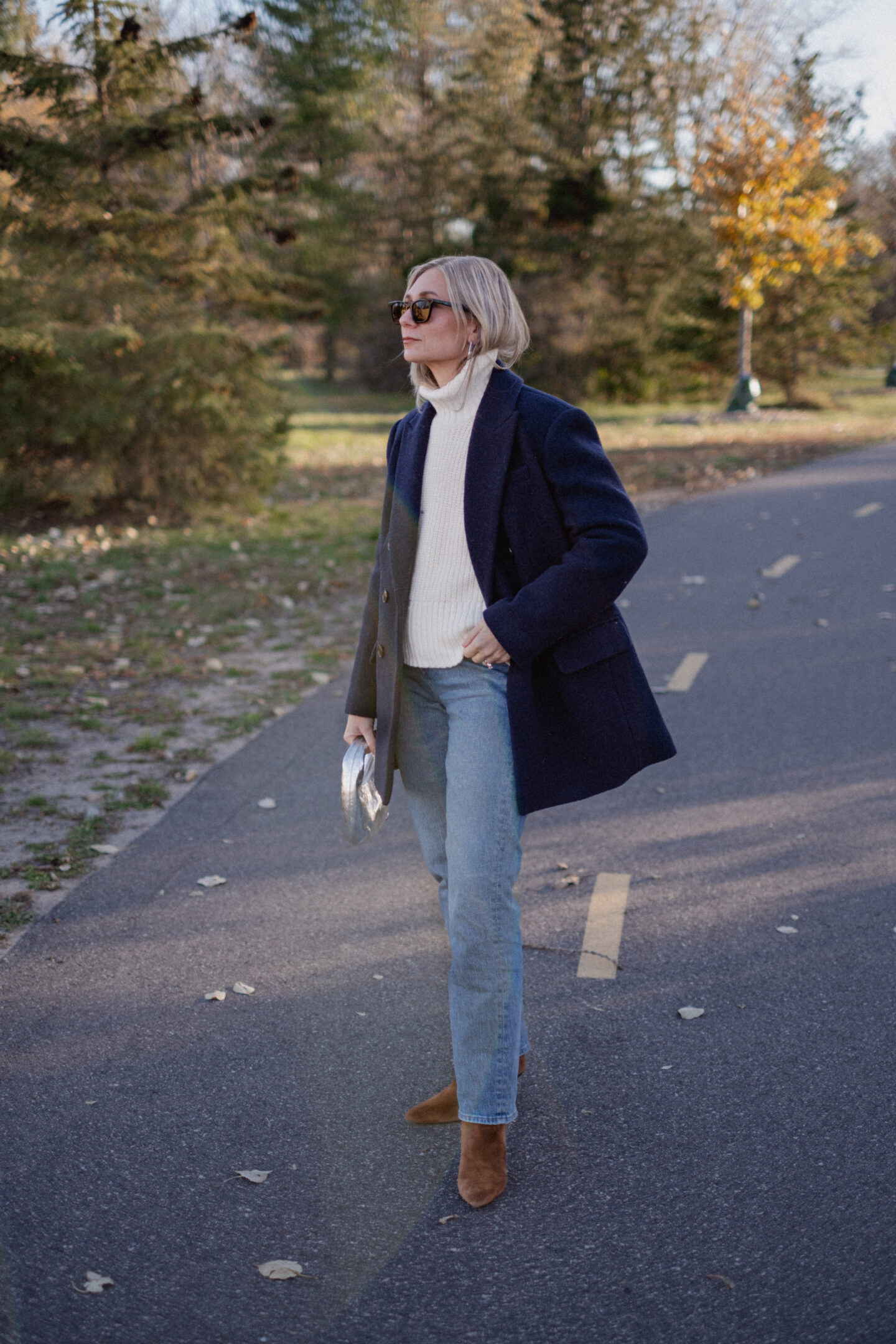 Karin Emily stands on a pathway wearing a chunky, ribbed cream turtleneck sweater under a navy wool coat with a pair of mid wash blue straight leg jeans, a pair of pointed, suede booties and a silver handbag