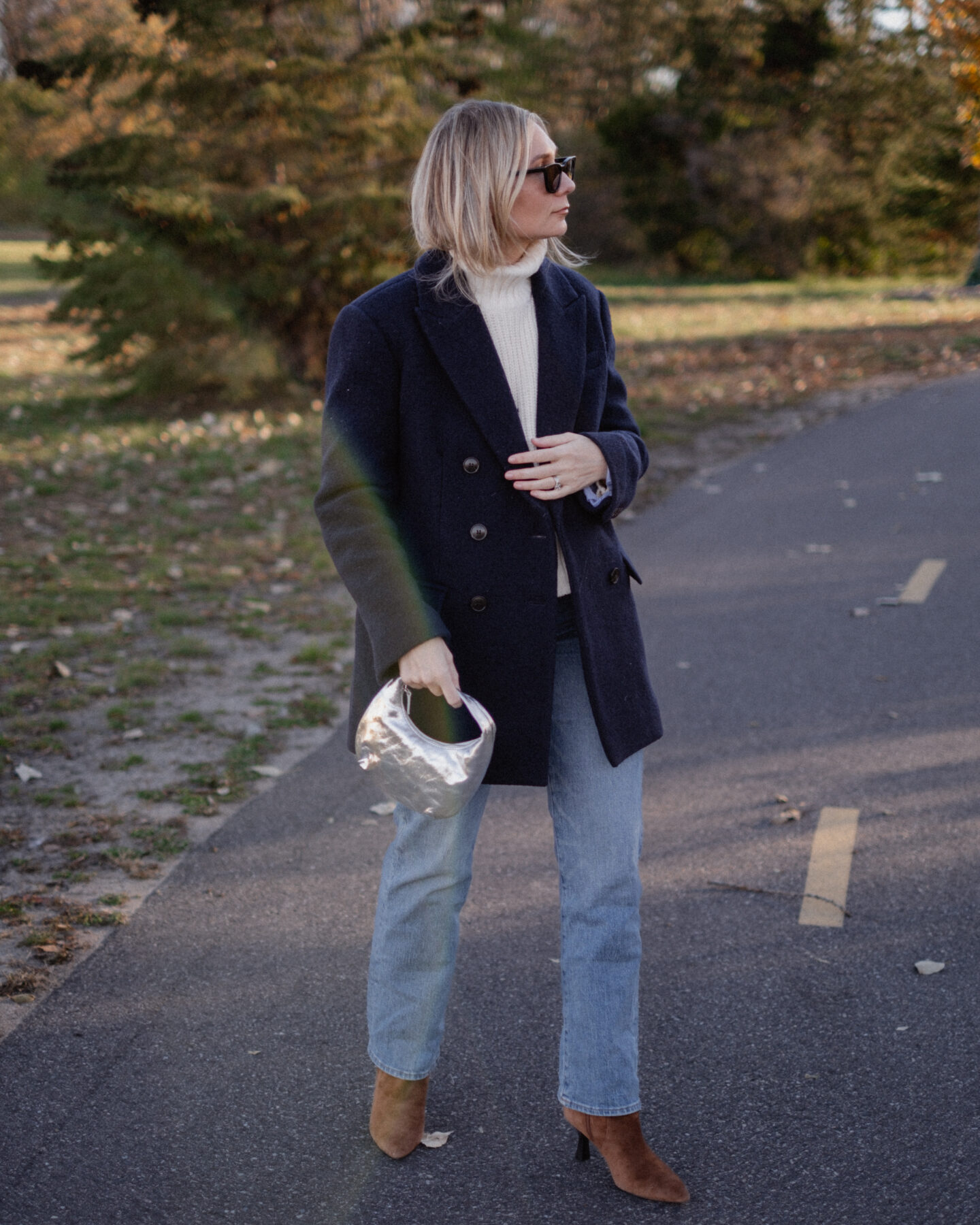 Karin Emily stands on a pathway wearing a chunky, ribbed cream turtleneck sweater under a navy wool coat with a pair of mid wash blue straight leg jeans, a pair of pointed, suede booties and a silver handbag