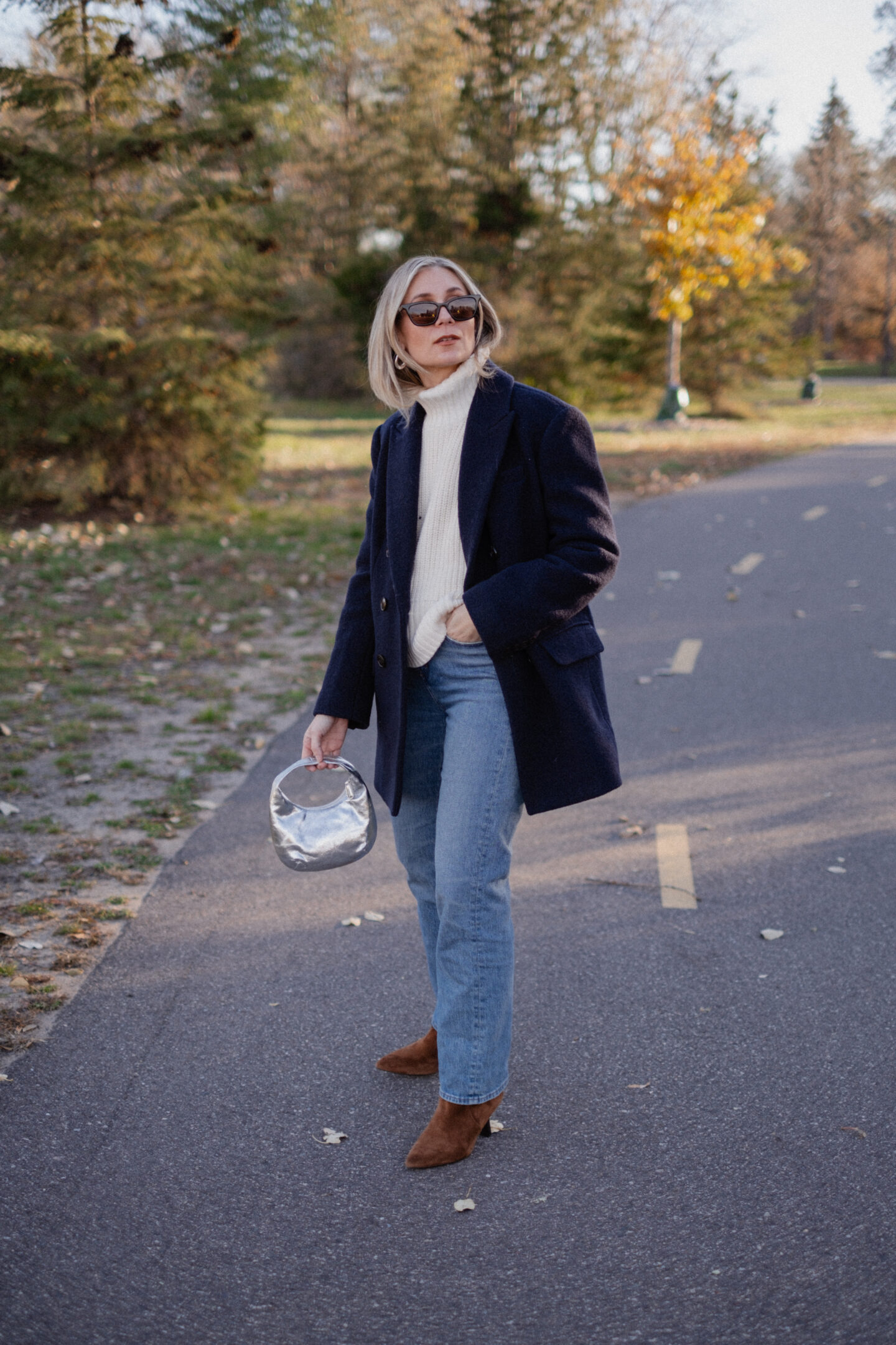 Karin Emily stands on a pathway wearing a chunky, ribbed cream turtleneck sweater under a navy wool coat with a pair of mid wash blue straight leg jeans, a pair of pointed, suede booties and a silver handbag