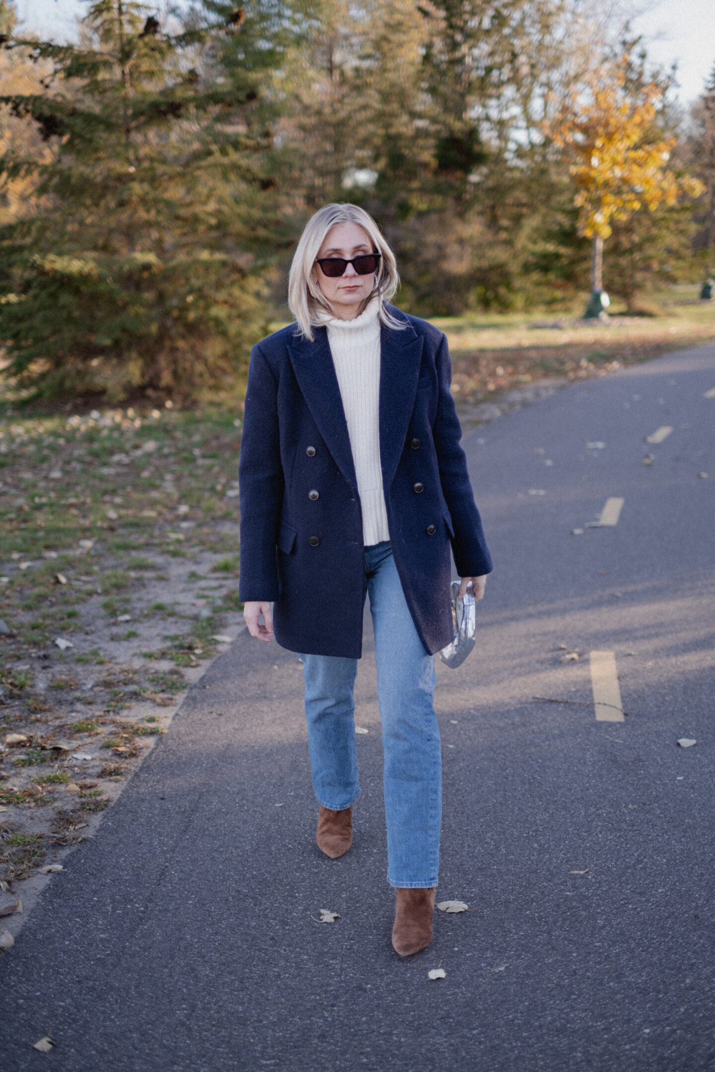 Karin Emily stands on a pathway wearing a chunky, ribbed cream turtleneck sweater under a navy wool coat with a pair of mid wash blue straight leg jeans, a pair of pointed, suede booties and a silver handbag