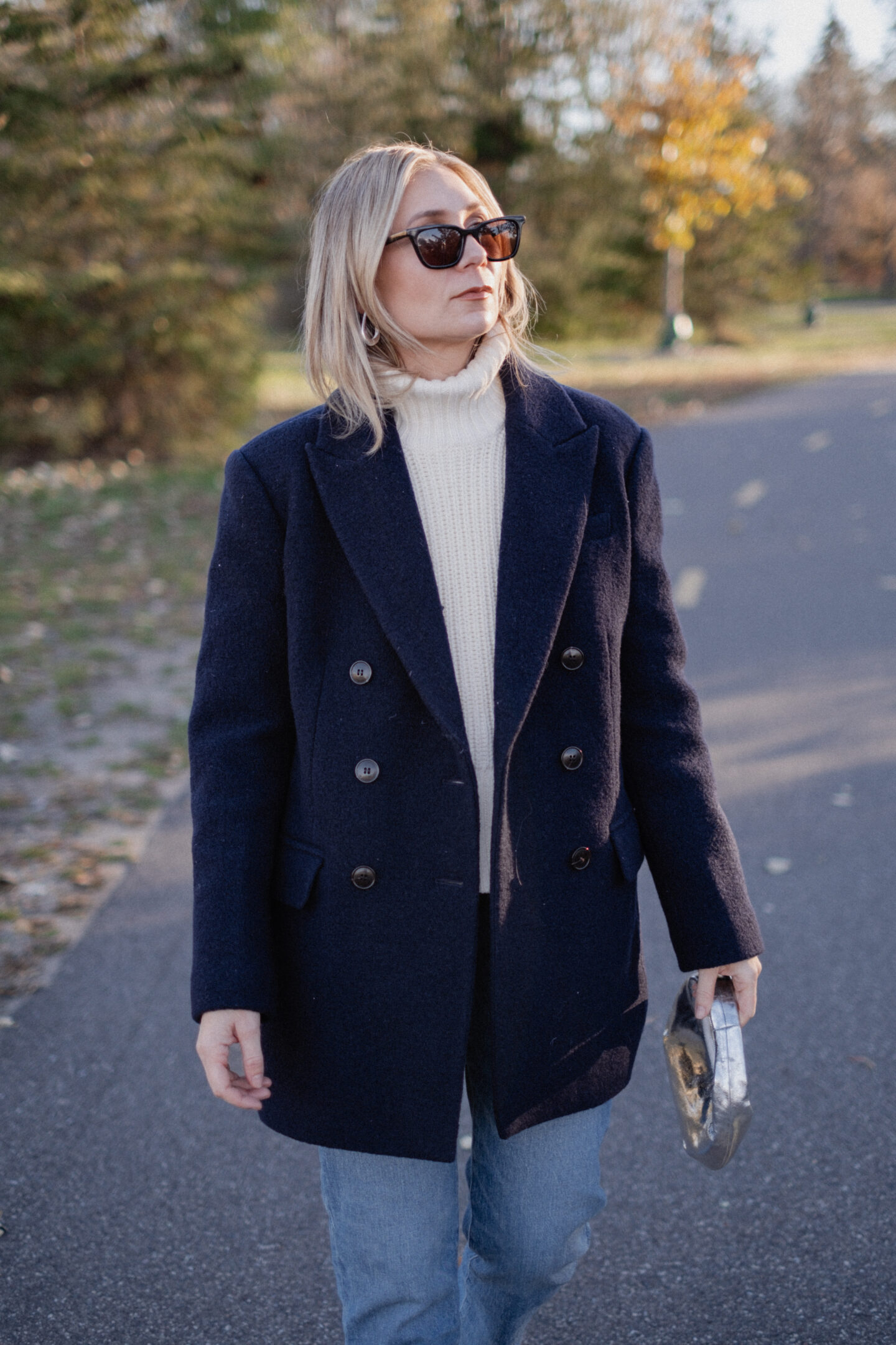 Karin Emily stands on a pathway wearing a chunky, ribbed cream turtleneck sweater under a navy wool coat with a pair of mid wash blue straight leg jeans, a pair of pointed, suede booties and a silver handbag