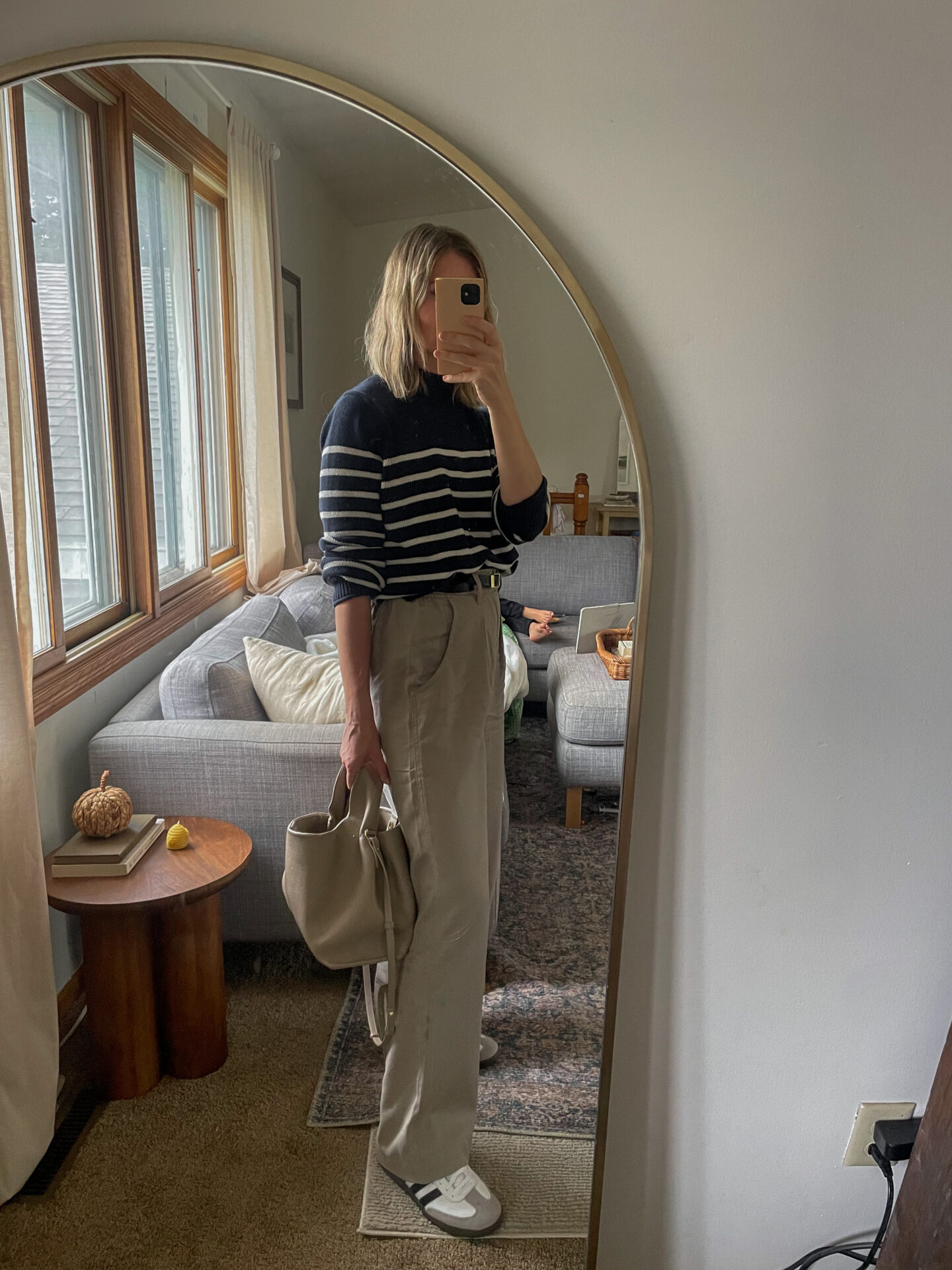 Karin Emily takes a mirror selfie in her room wearing a striped navy sweater with a pair of gray pleated pants and adidas samba sneakers and all are on sale for Black Friday Sales