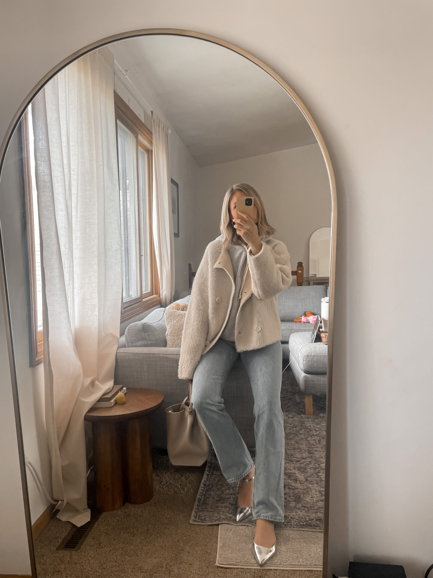 Karin Emily takes a mirror selfie in her living room wearing  a faux fur jacket over a gray sweatshirt with light wash jeans and silver flats