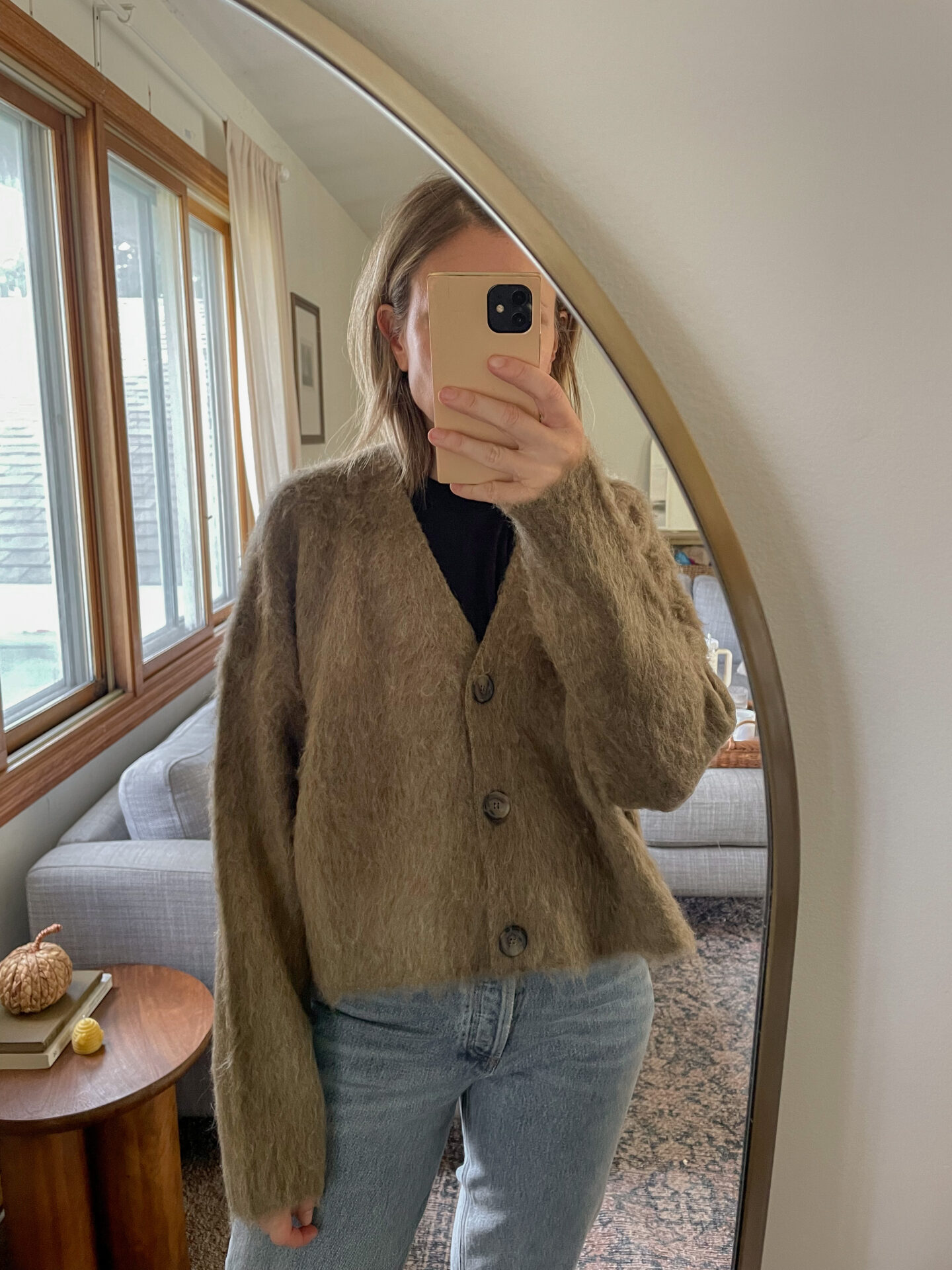 Karin Emily takes a mirror selfie in her living room wearing a fuzzy cardigan with light wash jeans