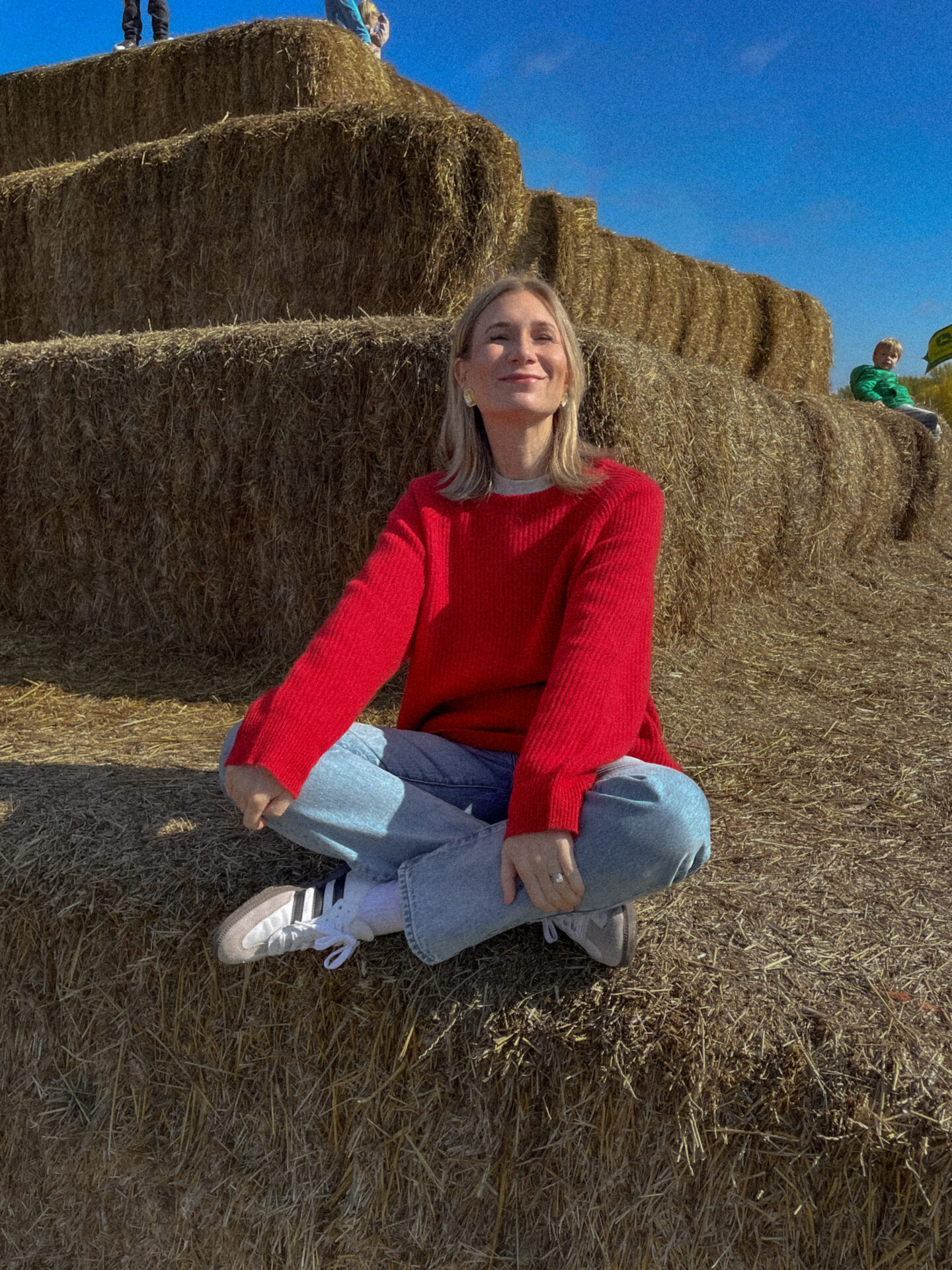 Karin Emily sits on a stack of hay bales wearing a red cashmere sweater with light wash jeans and adidas gazelle sneakers