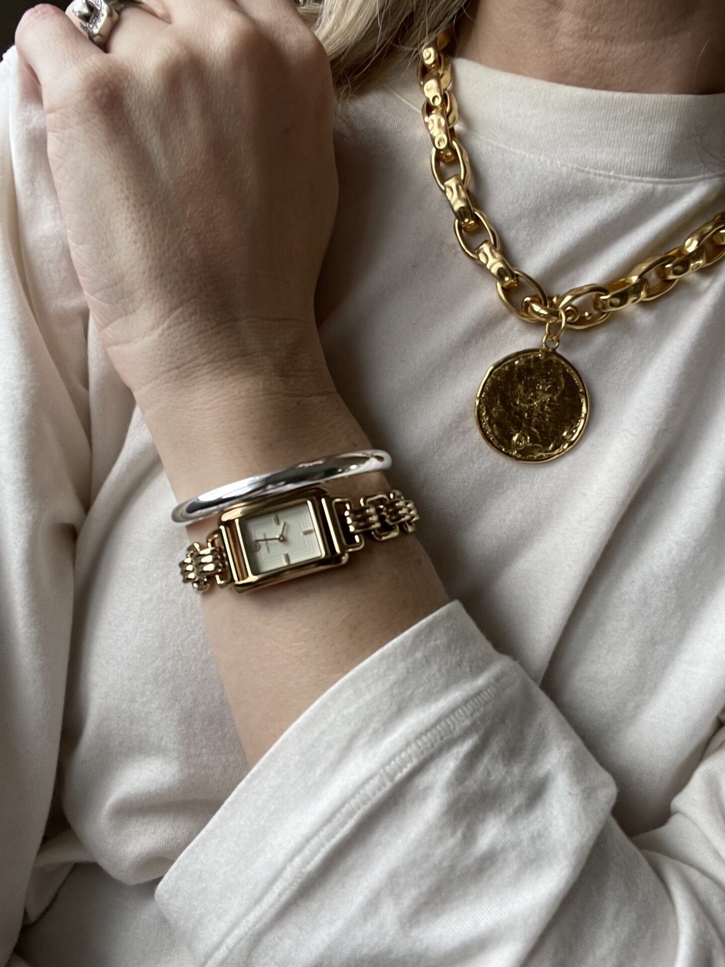 Karin Emily shows her jewelry that includes a gold watch with a silver bangle and a chunky gold chain necklace with a gold circle medallion 