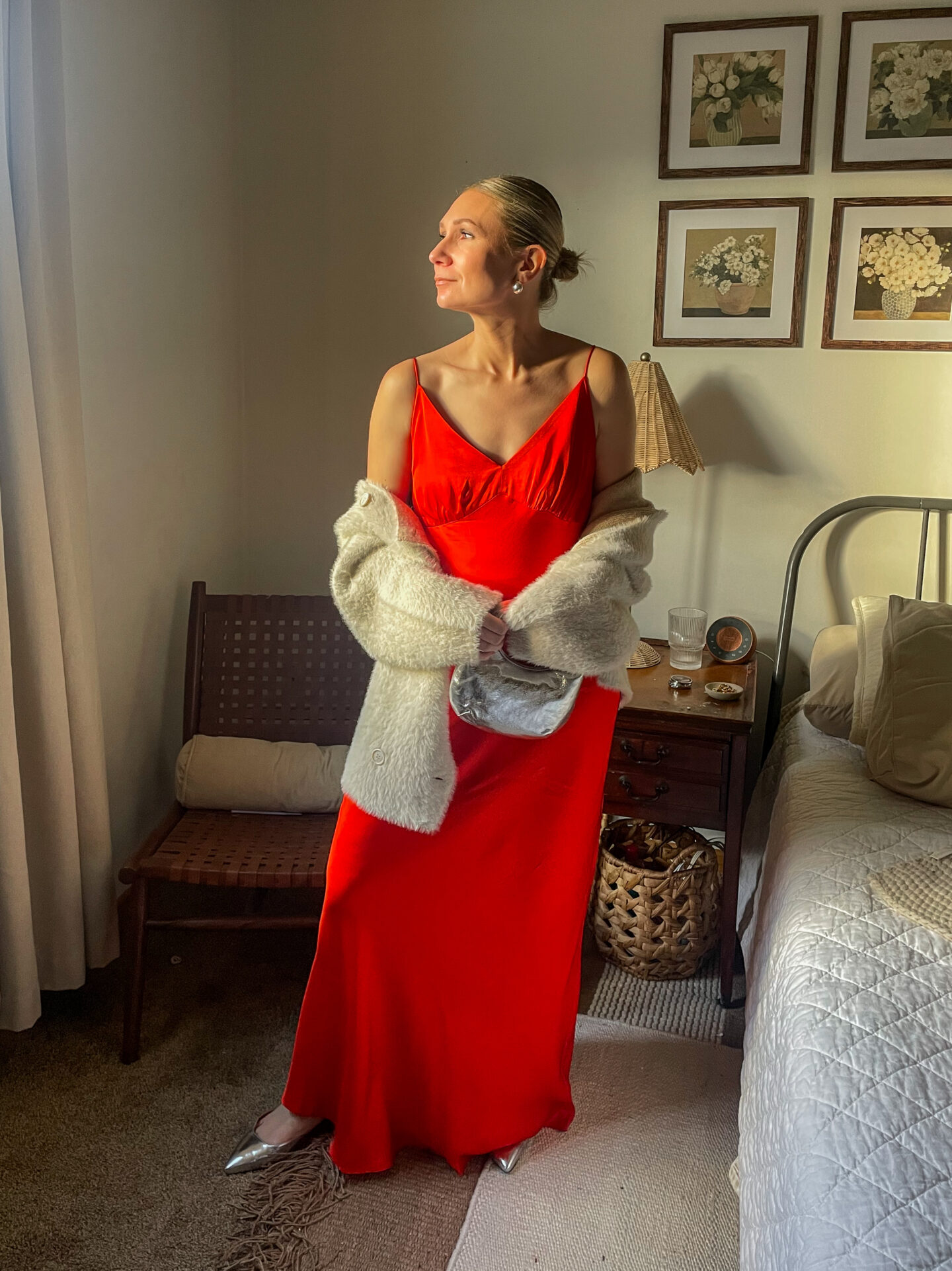 Karin Emily stands in her room wearing a bright red maxi slip dress with a pair of silver shoes and a faux fur drape coat