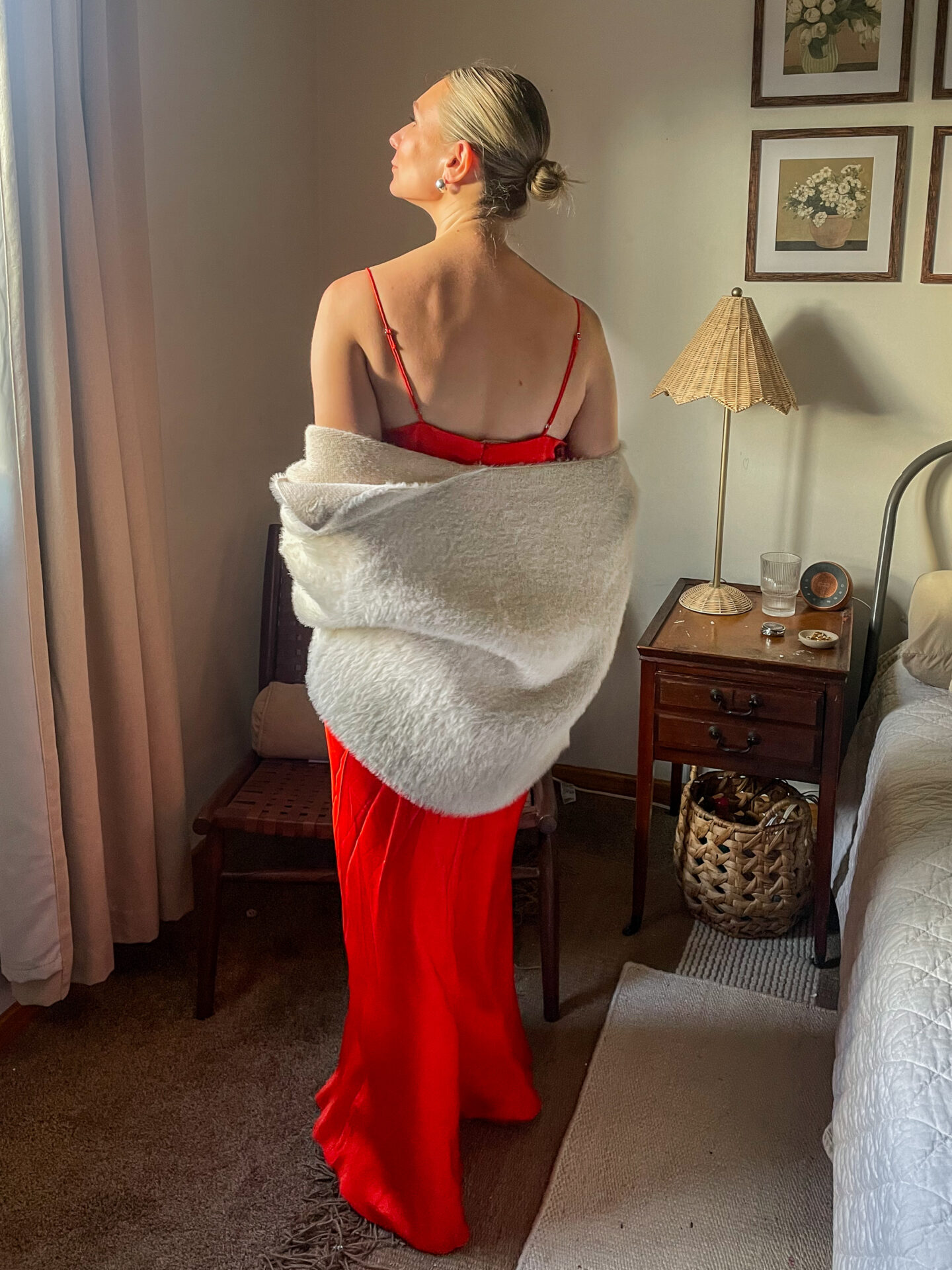 Karin Emily stands in her room wearing a bright red maxi slip dress with a pair of silver shoes and a faux fur drape coat